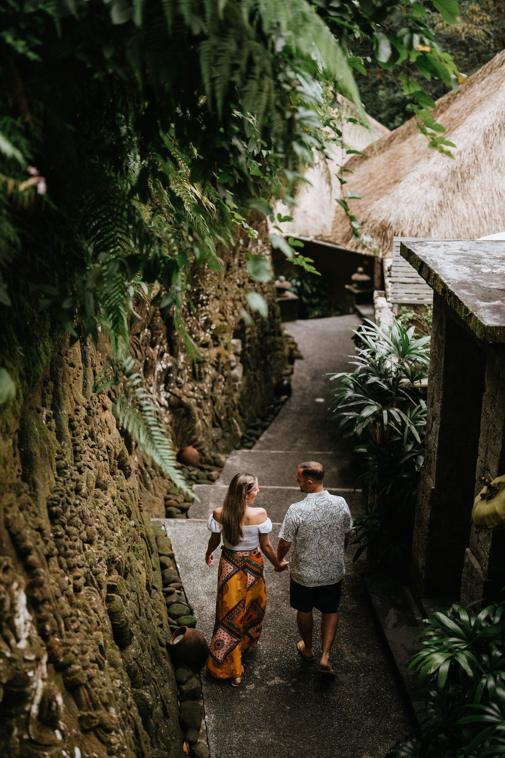 Two people walk down stone steps surrounded by tropical plants and rock walls.