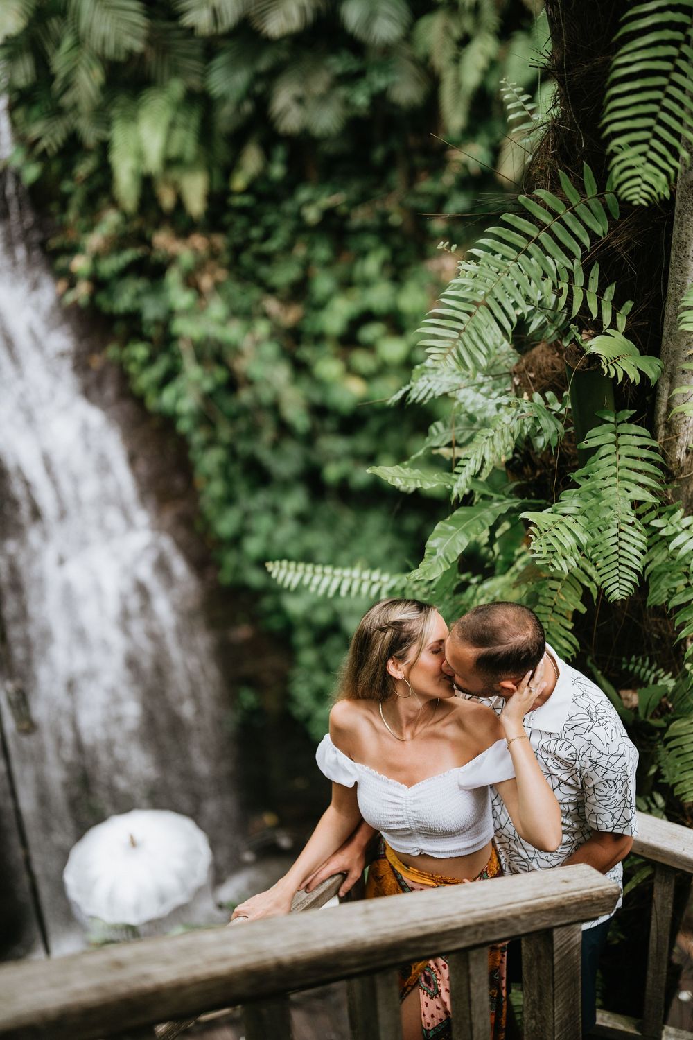 A tender embrace photographed near a waterfall with ferns and tropical foliage.