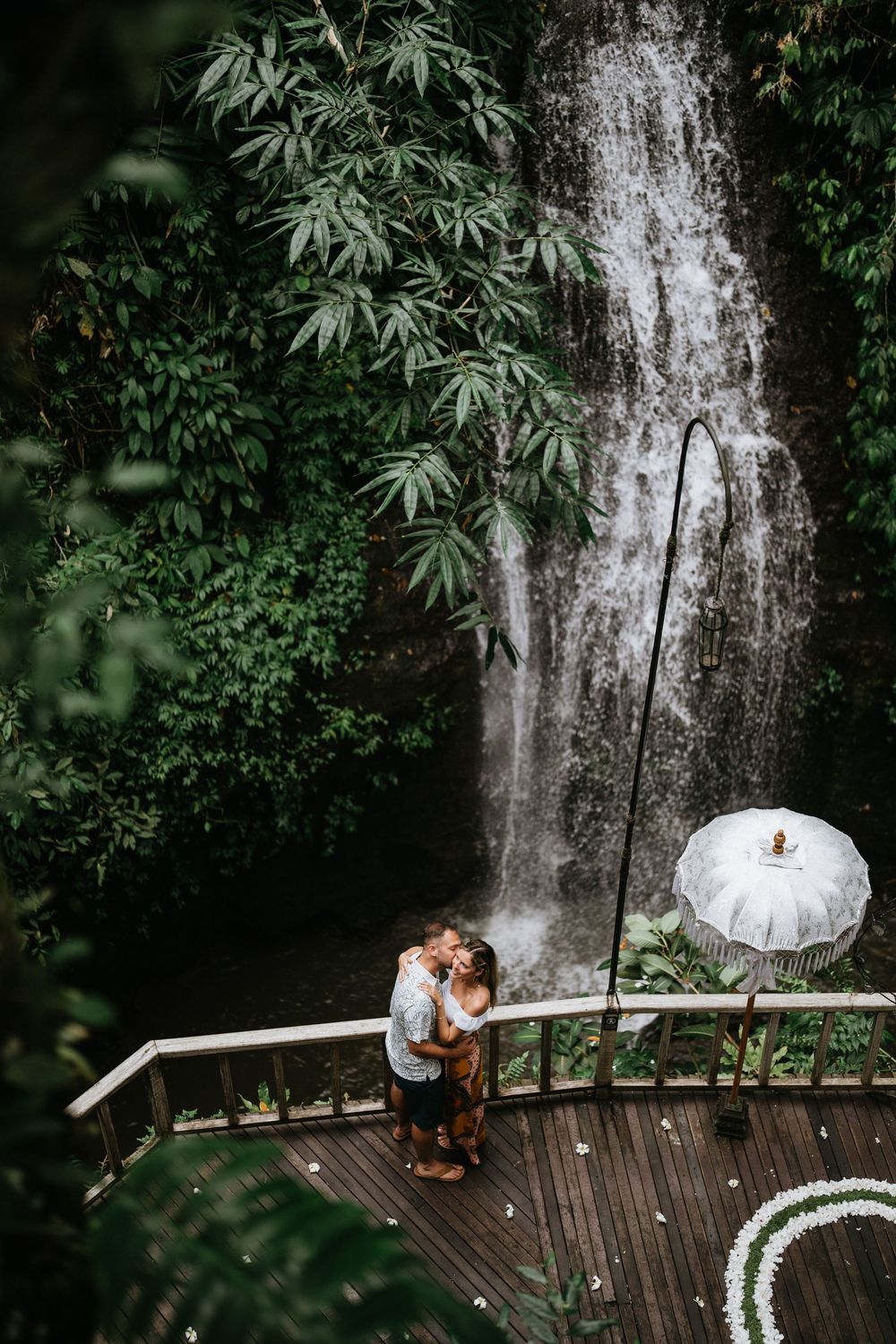 Couple sharing intimate moment on wooden deck with waterfall backdrop.