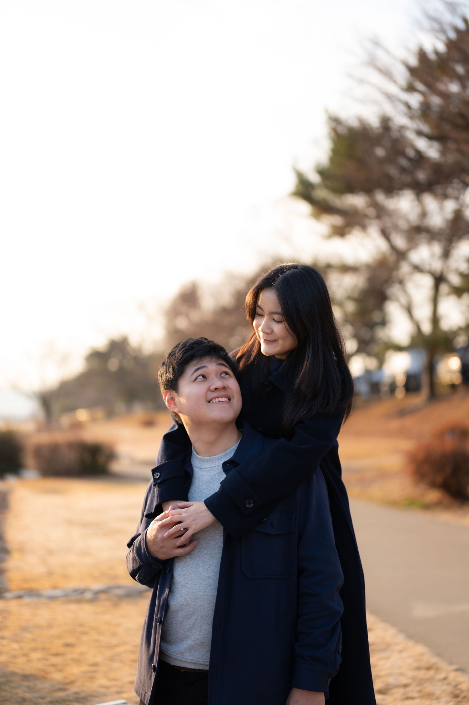 Smiling couple embracing outdoors at golden hour, woman hugging man from behind on park path.
