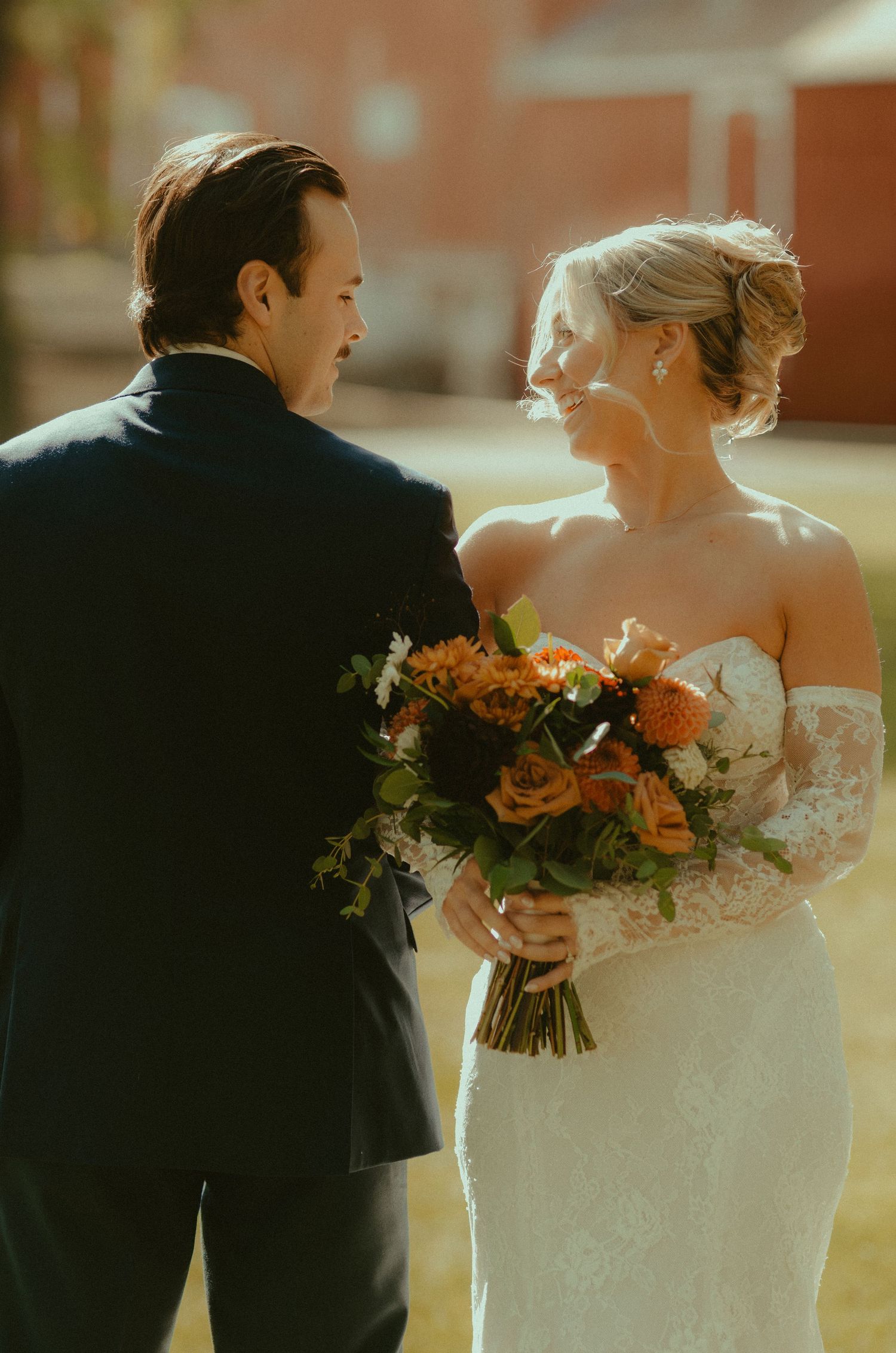 Bride in lace gown holds orange bouquet, smiling at groom in navy suit during outdoor wedding ceremony.