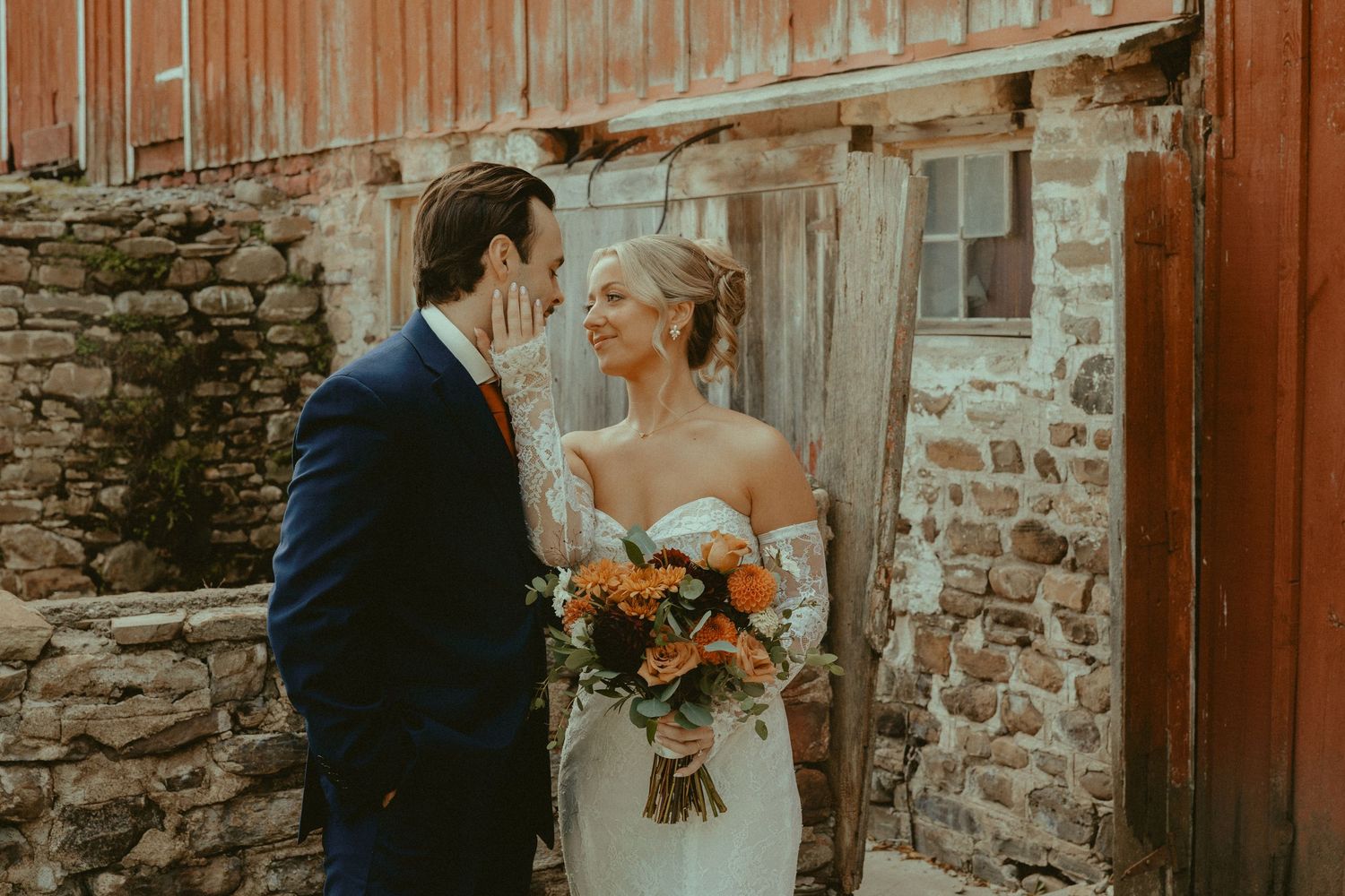 Bride and groom share a romantic moment outside a rustic stone barn, bride holding vibrant orange fall bouquet.