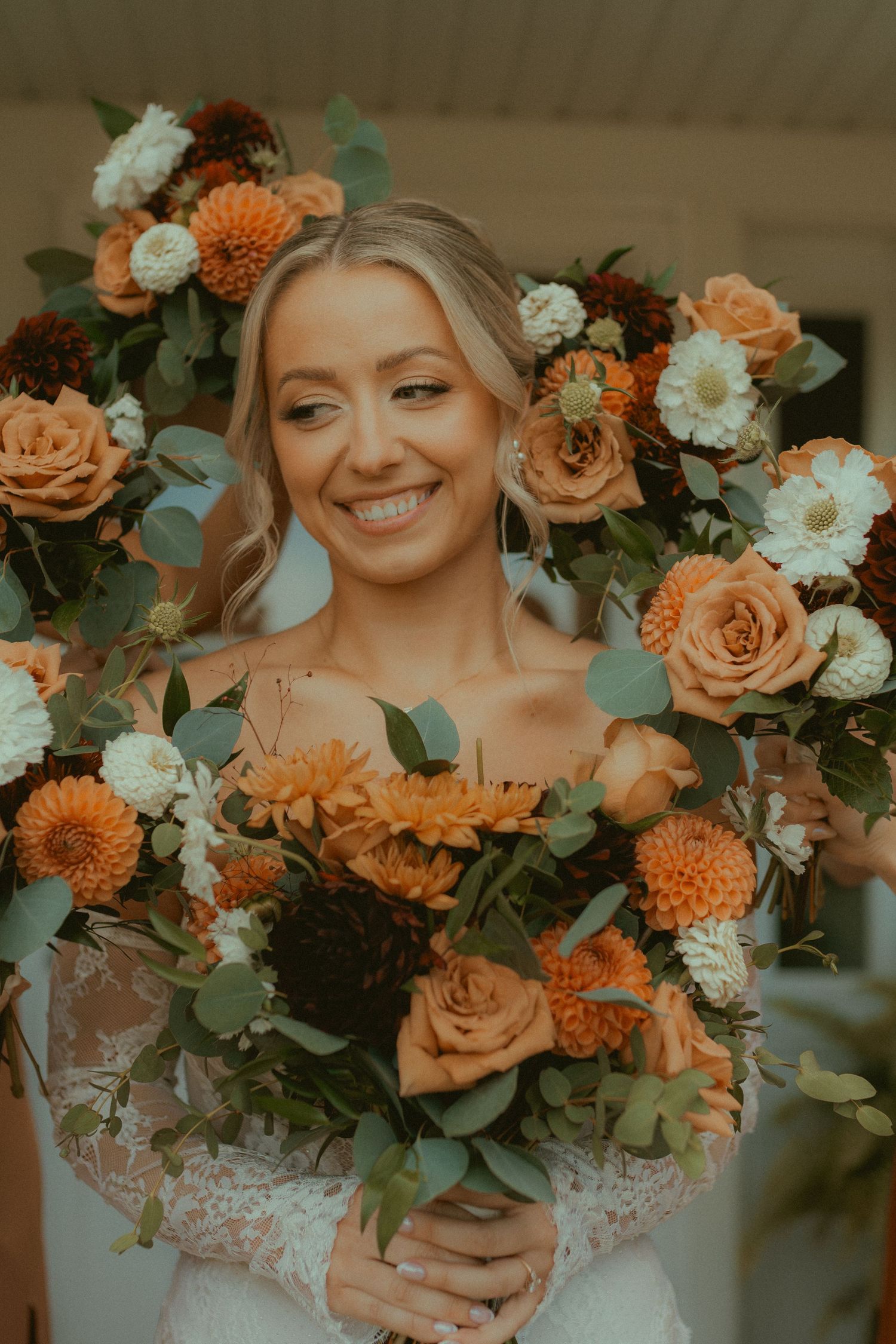 Smiling bride in lace gown holds orange, white, and burgundy floral bouquet with eucalyptus greenery.