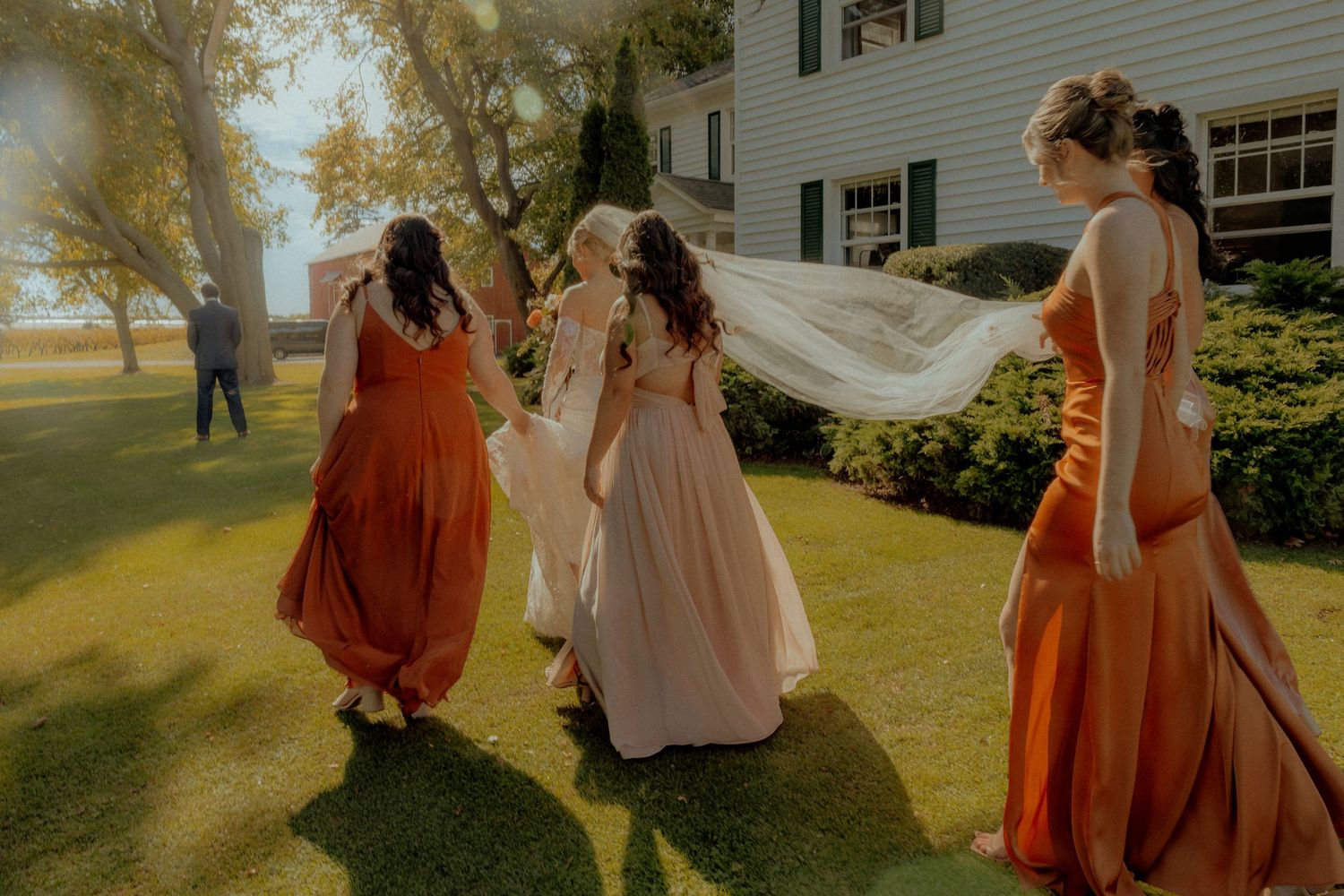 Bridesmaids in rust and blush gowns walk with bride whose veil flows in the breeze on a sunny lawn.