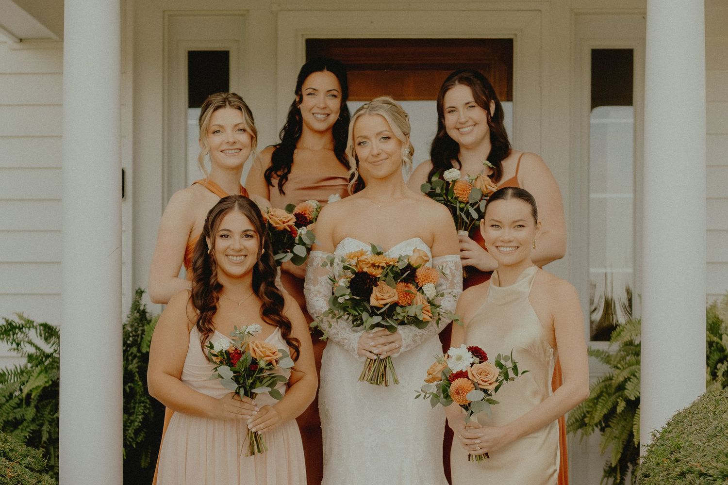 Bride in white gown poses with five bridesmaids in champagne dresses holding colorful bouquets on white porch steps.