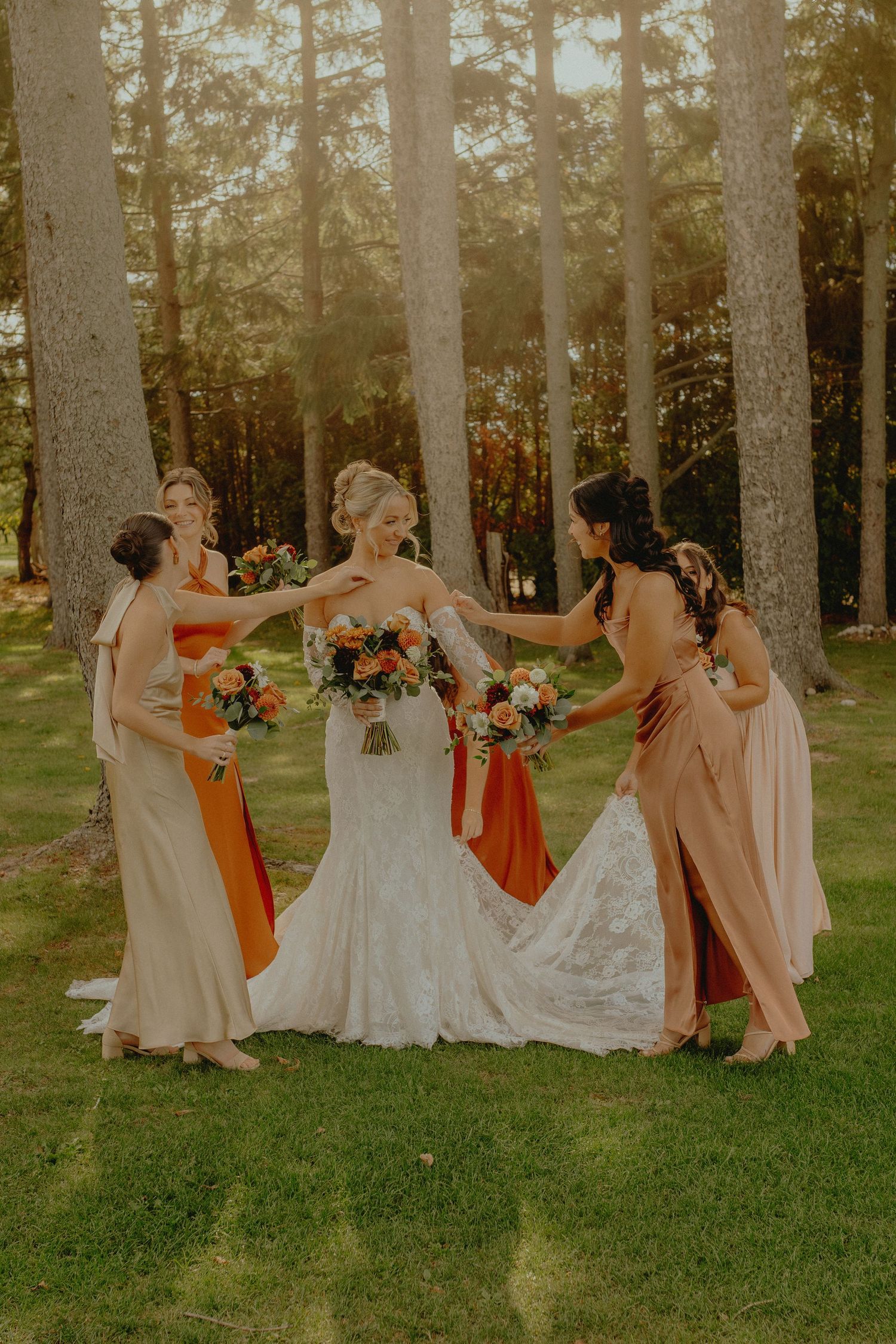 Bride in lace gown surrounded by bridesmaids in orange and blush dresses holding bouquets in a sunlit forest setting.