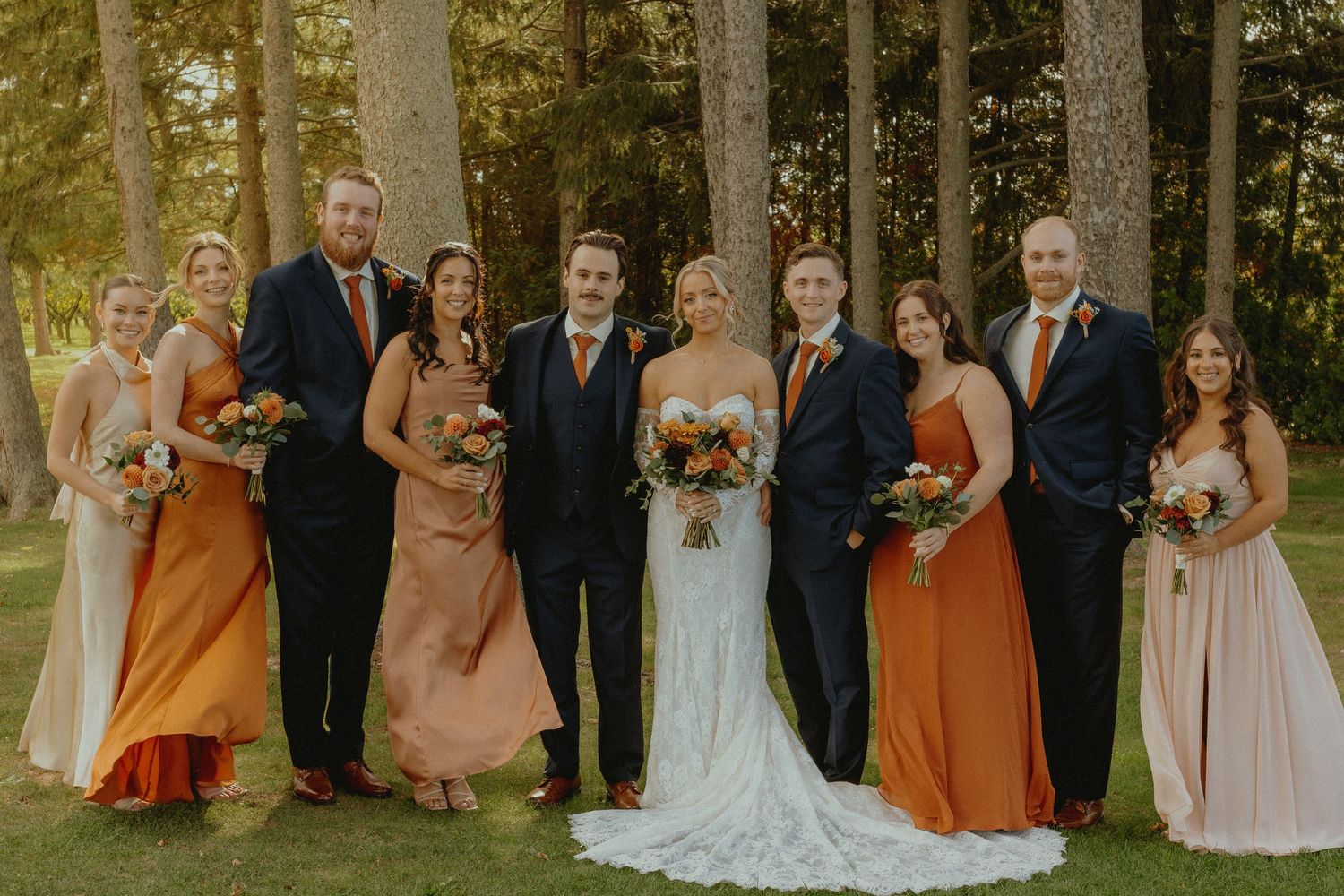 Wedding party poses outdoors among tall trees, bridesmaids in rust and blush gowns, groomsmen in navy suits.