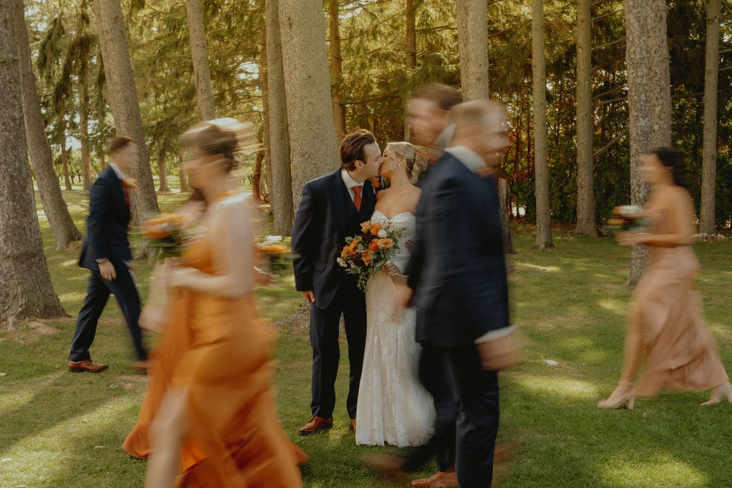 Bride and groom share a kiss surrounded by wedding party in motion blur among tall trees outdoors.