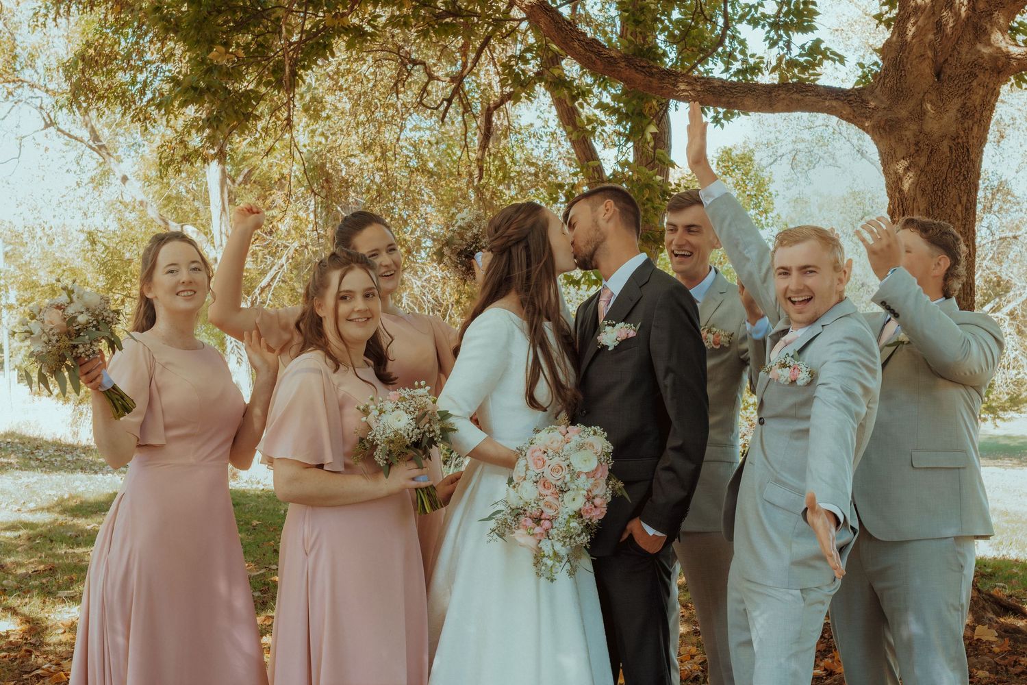 Wedding party in blush pink and gray attire gathers outdoors under trees for formal photos.