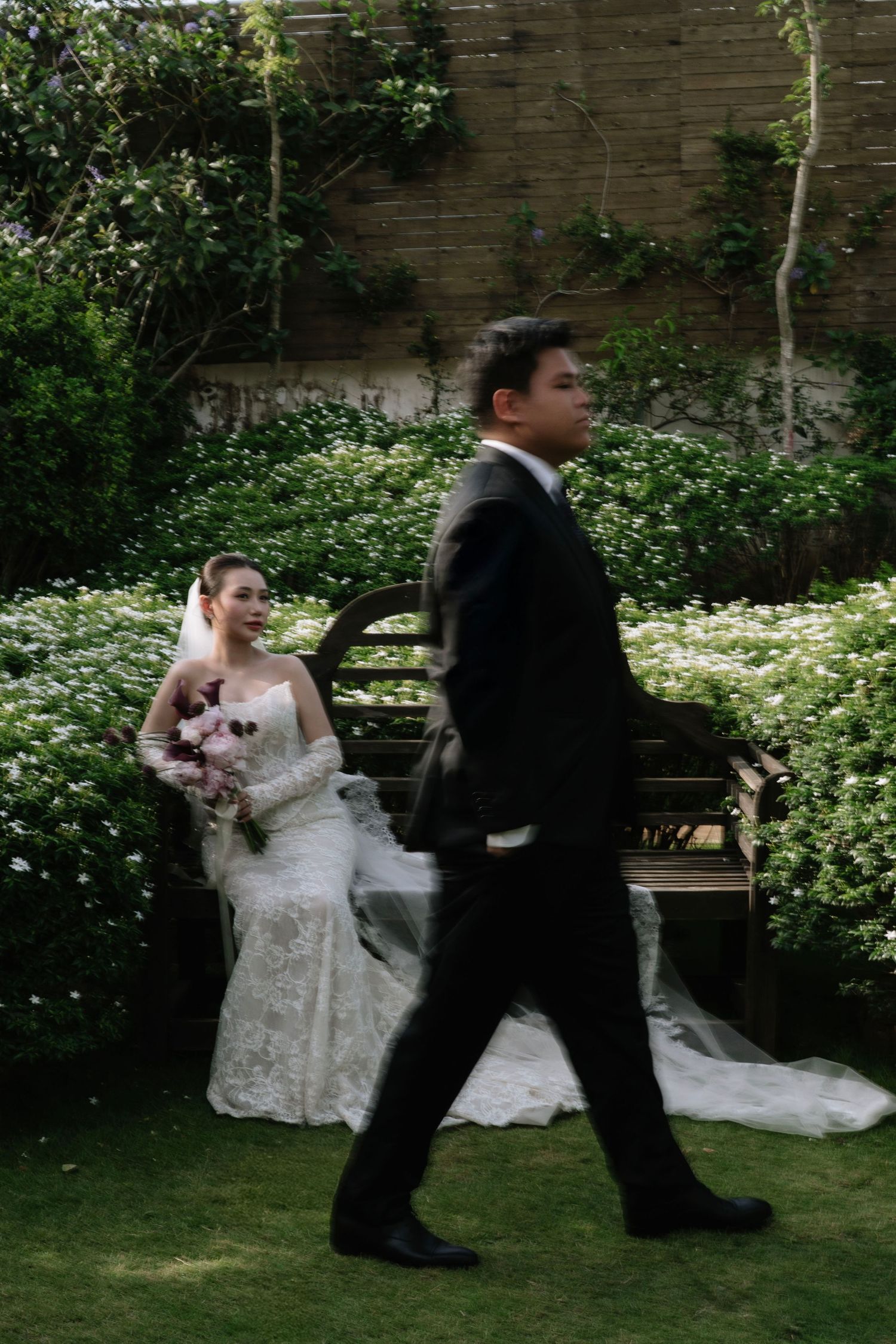 Bride in lace gown sitting on a garden bench holding a bouquet while groom walks past in a green outdoor setting.
