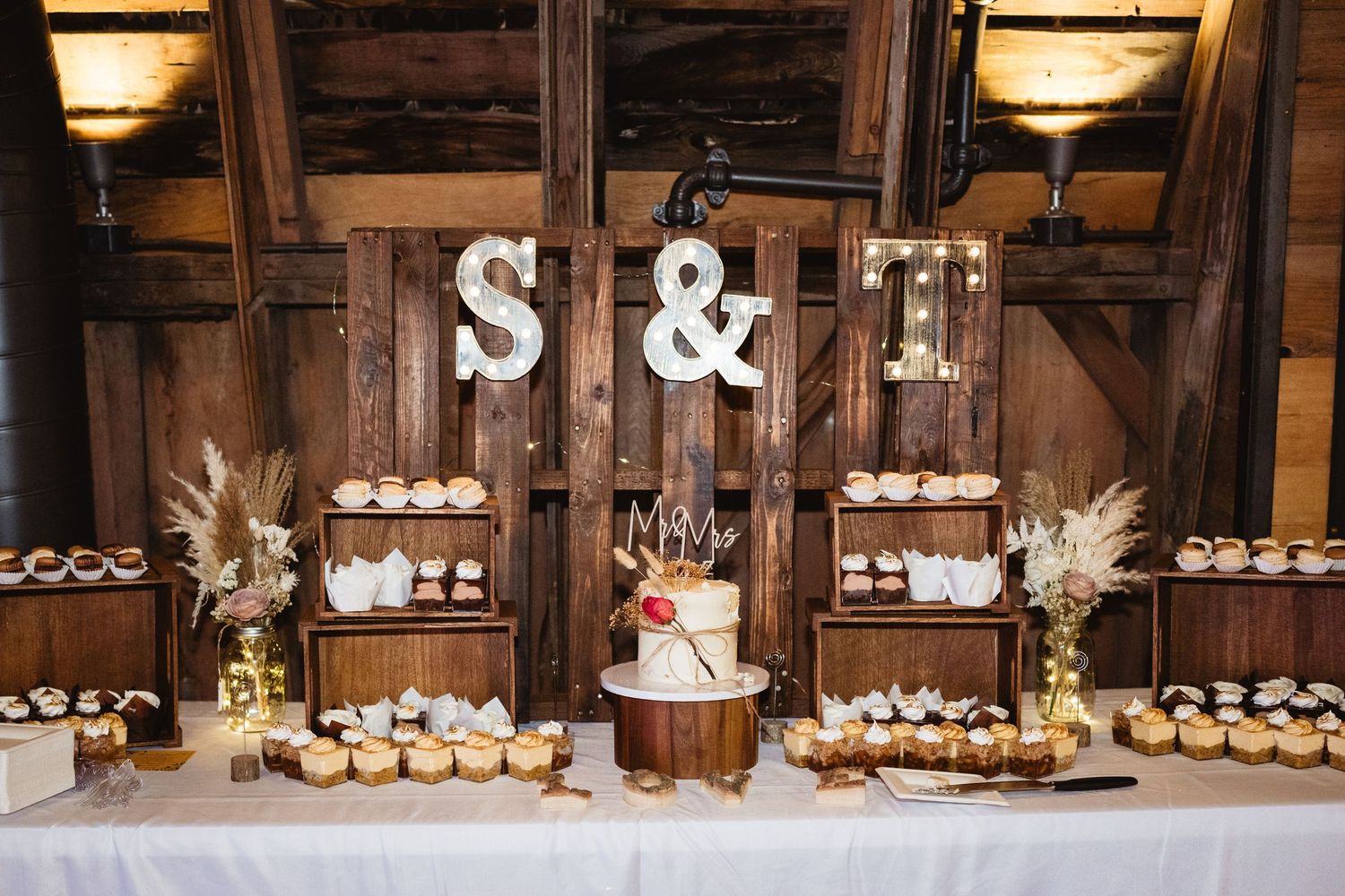 Rustic wedding dessert table with cupcakes and white cake displayed under wooden S&T letters in barn setting.