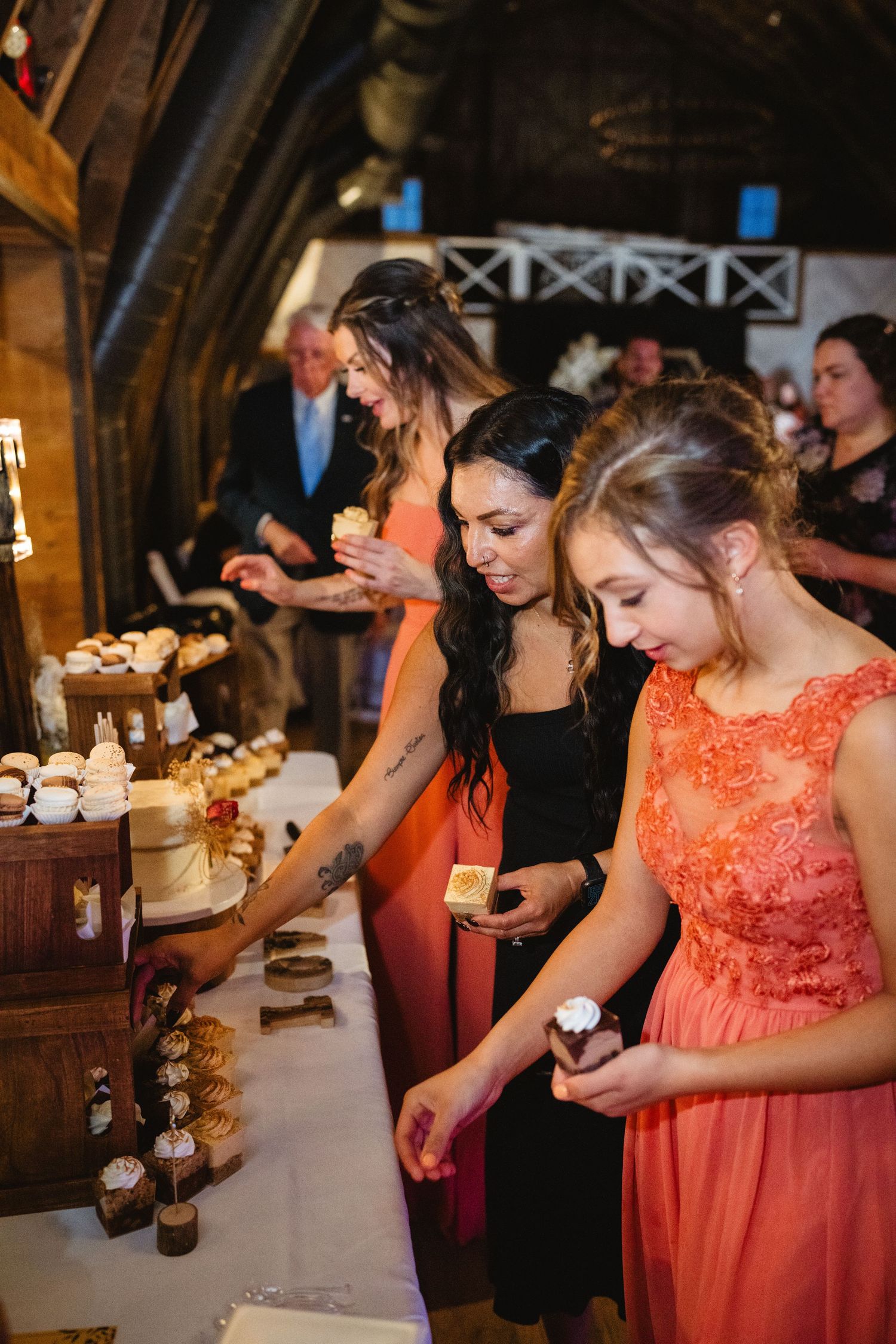 Wedding attendees in coral dresses and formal wear gather around a dessert table at a reception.