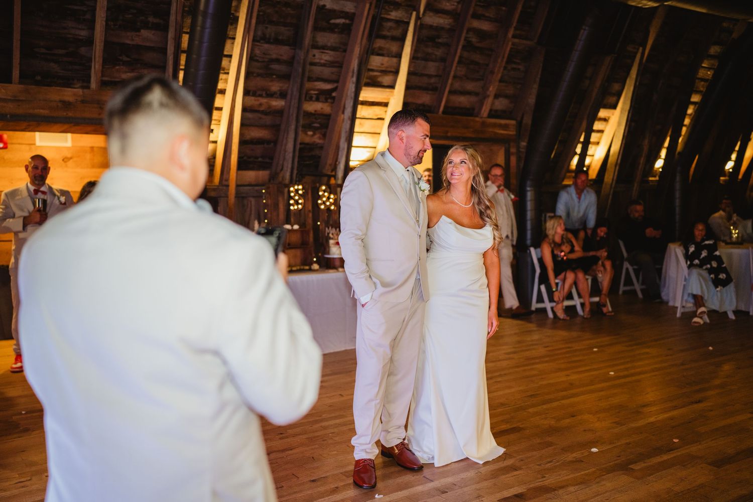People dance at a rustic wedding reception inside a warmly-lit barn venue with wooden beams and floors.