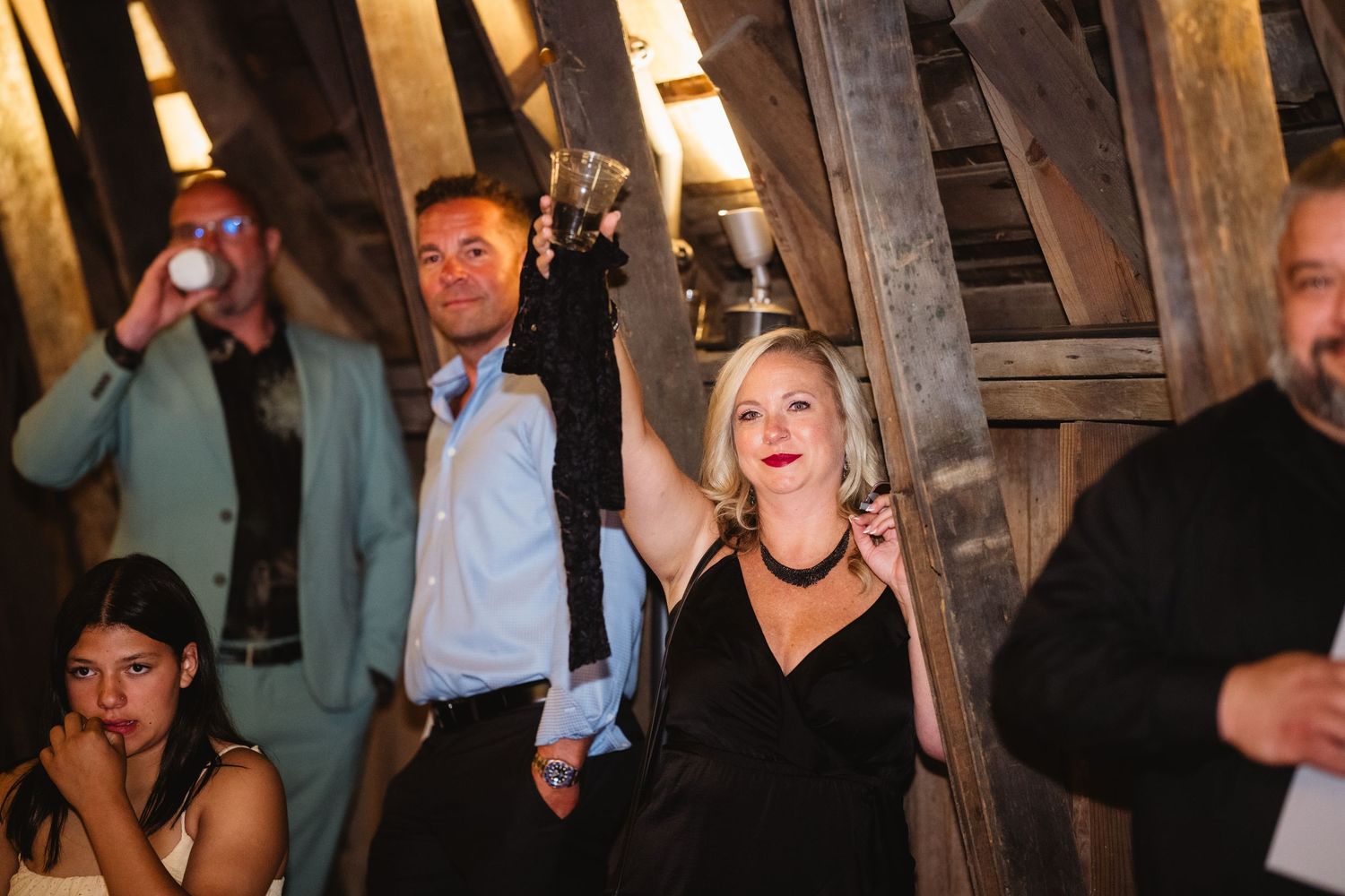 Wedding guests celebrate and dance under rustic wooden beams at an evening reception party.