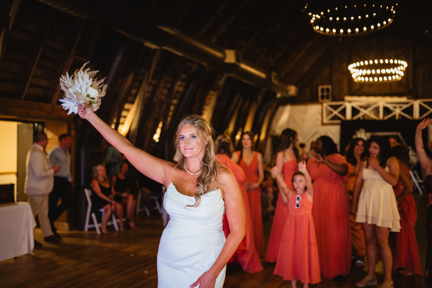 A bride raises her bouquet during a wedding reception in a rustic barn venue with string lights overhead.