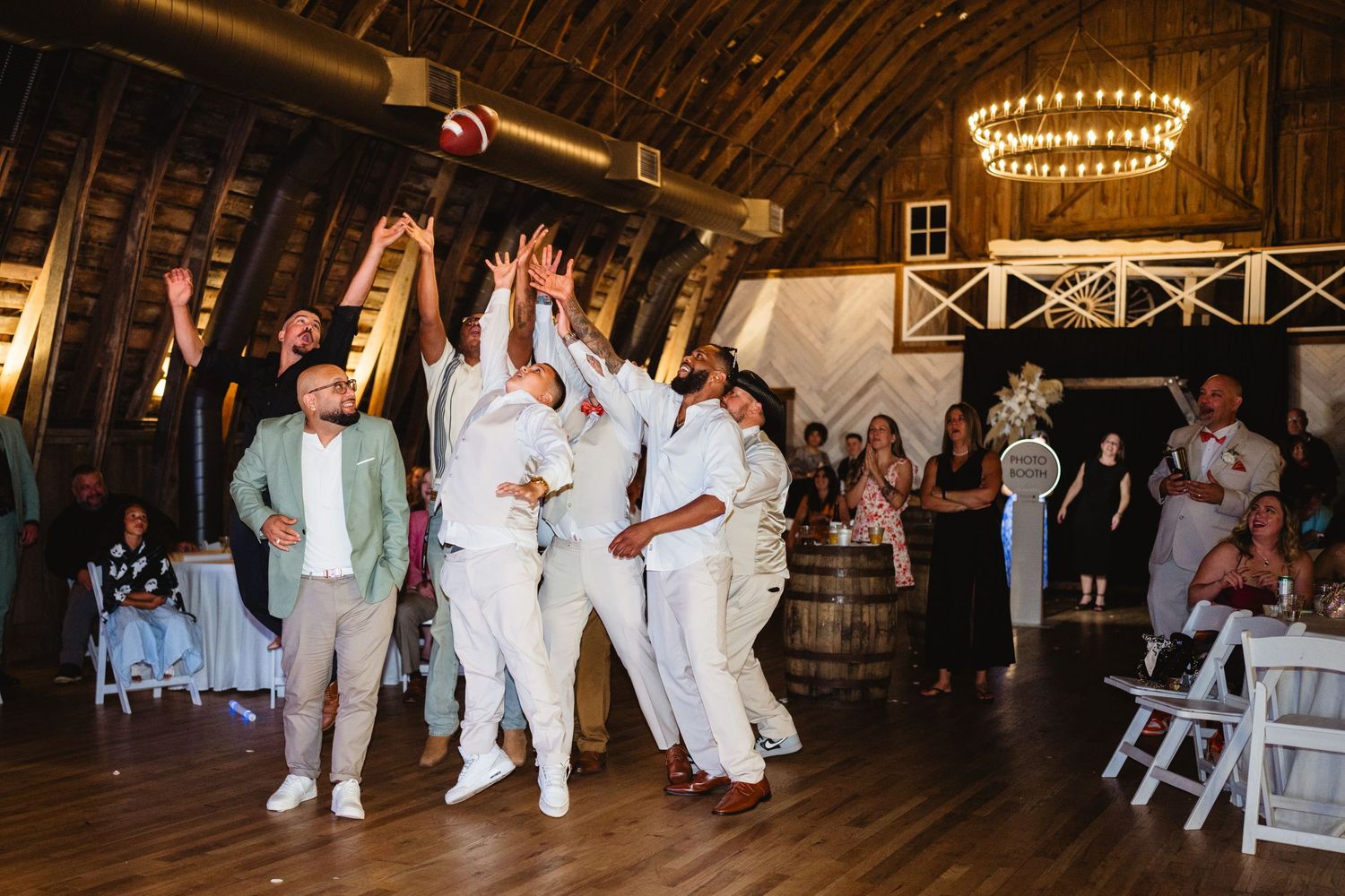 Wedding party members dancing during a reception celebration in a rustic barn venue illuminated by chandeliers.