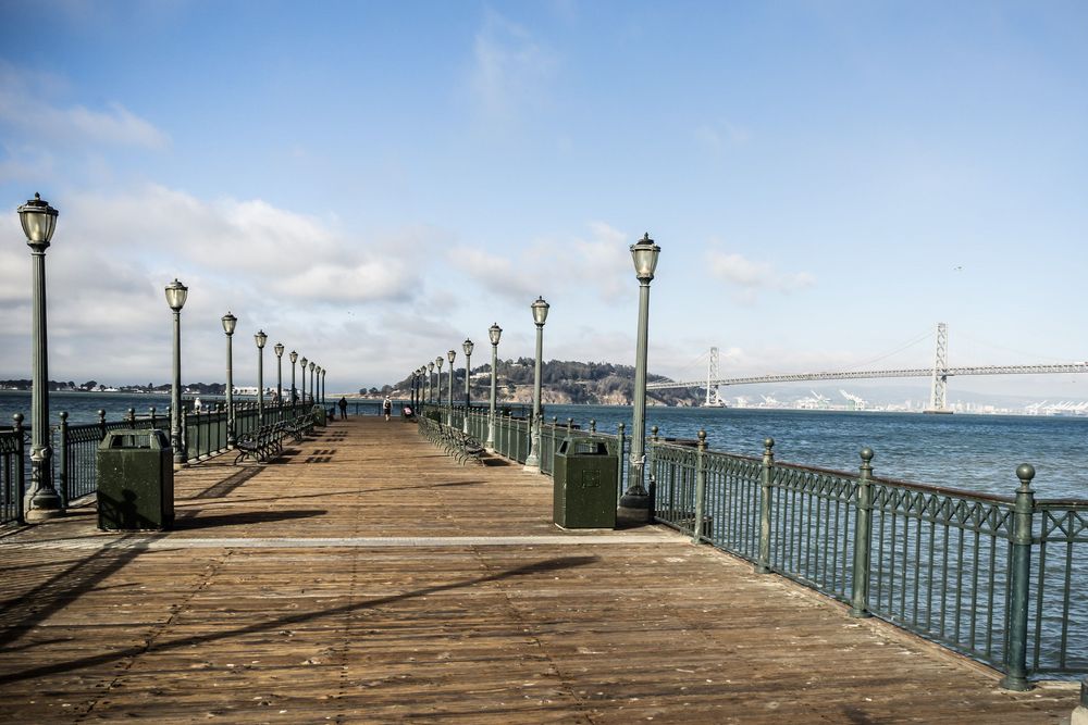 A long wooden pier with Victorian-style lampposts stretches into the blue bay under a partly cloudy sky.