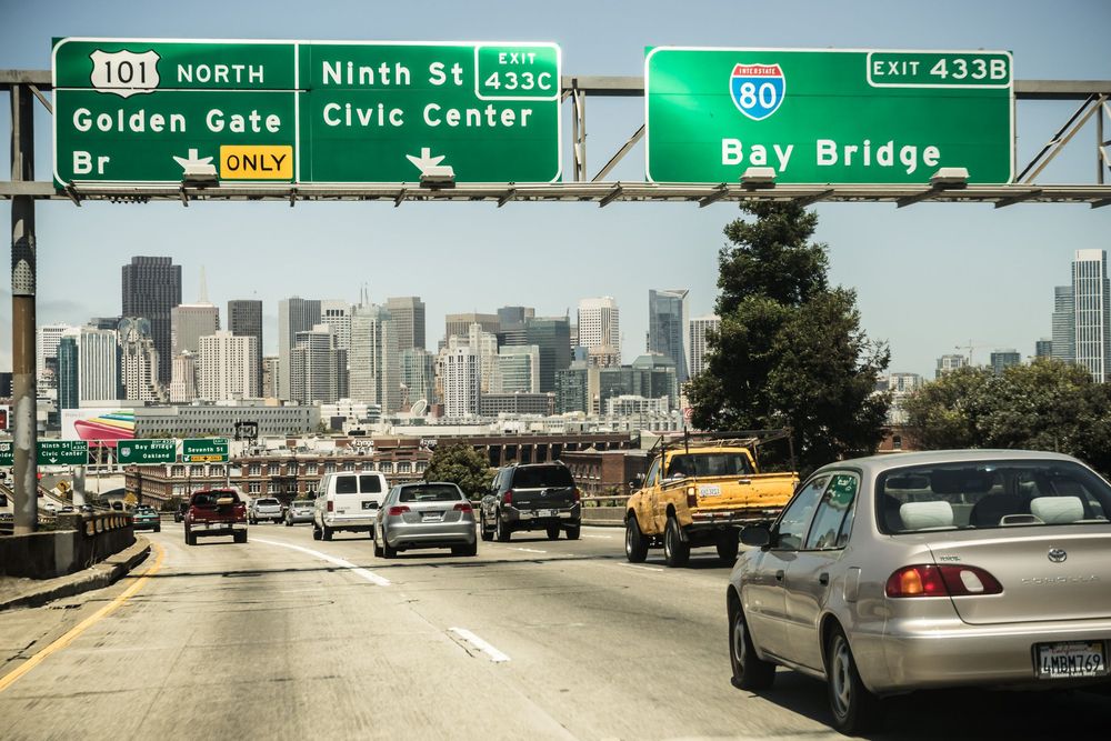Traffic on a San Francisco highway with road signs for Golden Gate and Bay Bridge against a downtown skyline view.