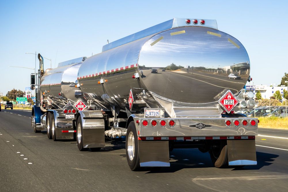 Six metallic tanker trucks line up on a curved highway against a blue sky with hills in the background.