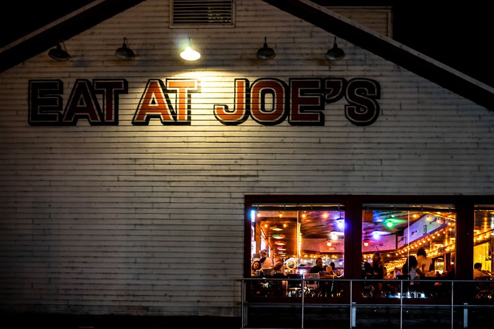 A neon-lit storefront of "Eat at Joe's" restaurant glows brightly in the nighttime darkness against a brick wall.