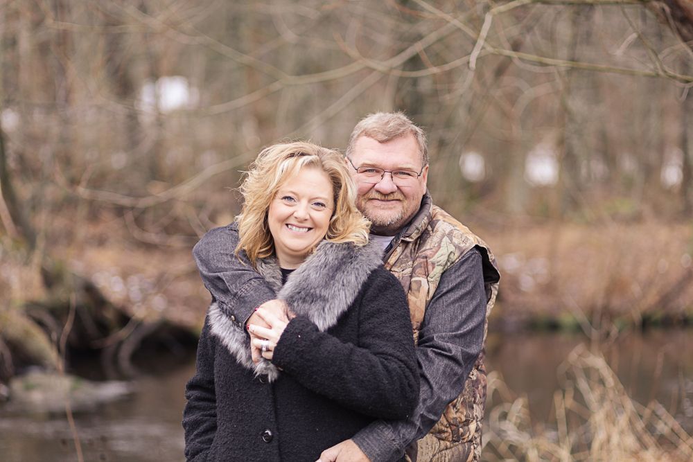 Couple embracing outdoors wearing winter coats near wooded area.