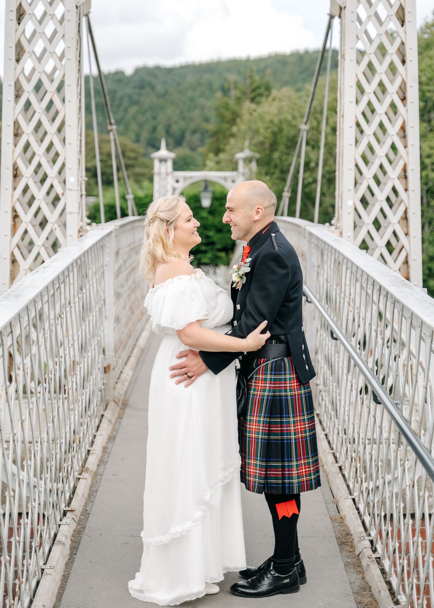 Romantic wedding couple embraces on suspension bridge with groom wearing traditional Scottish kilt.