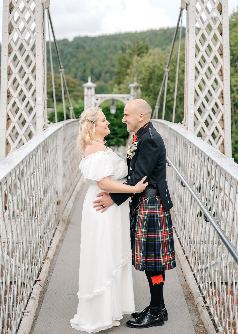 Romantic wedding couple embraces on suspension bridge with groom wearing traditional Scottish kilt.
