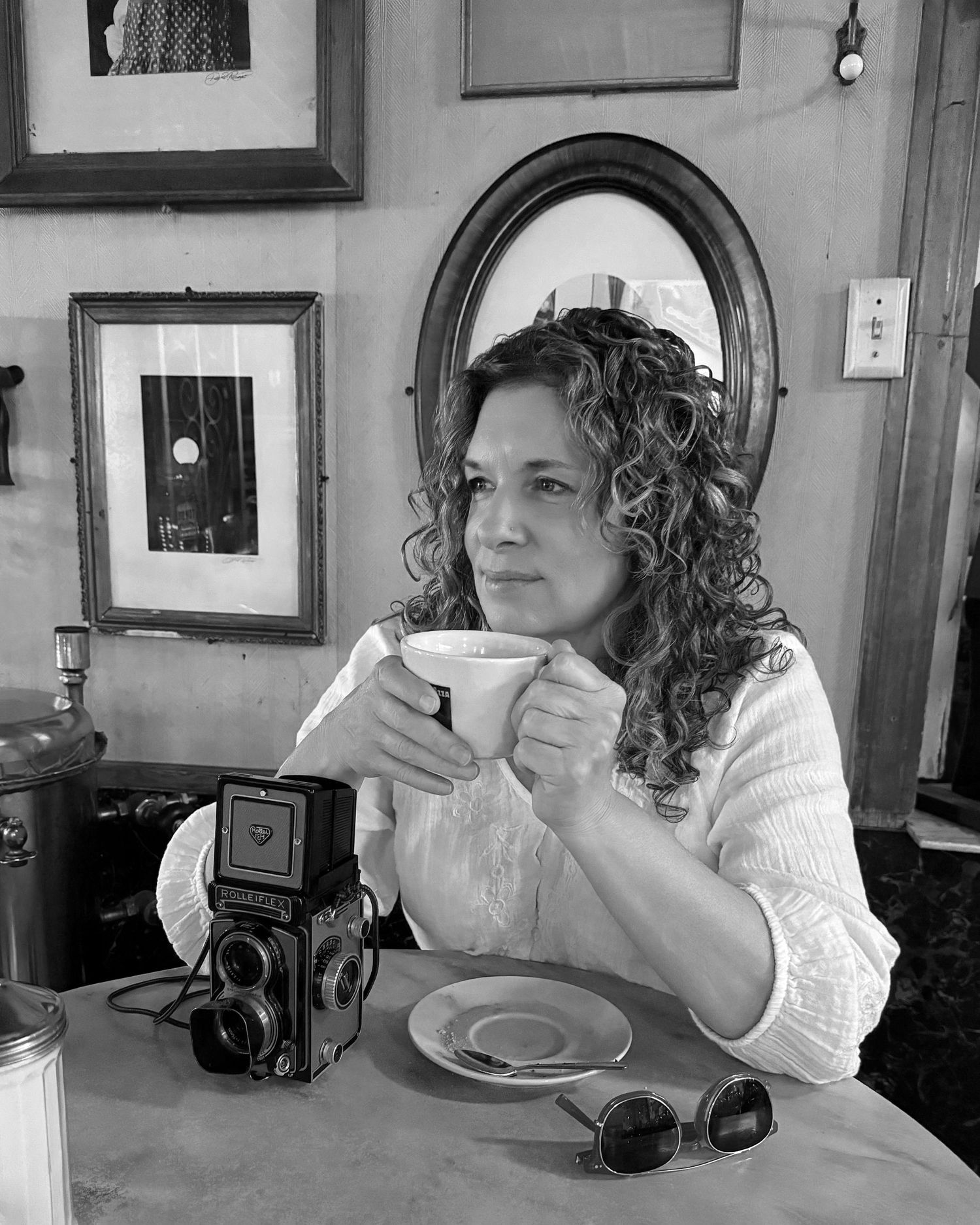 Black and white photo of a curly-haired woman holding a coffee cup beside a vintage twin-lens reflex camera in a cozy cafe.