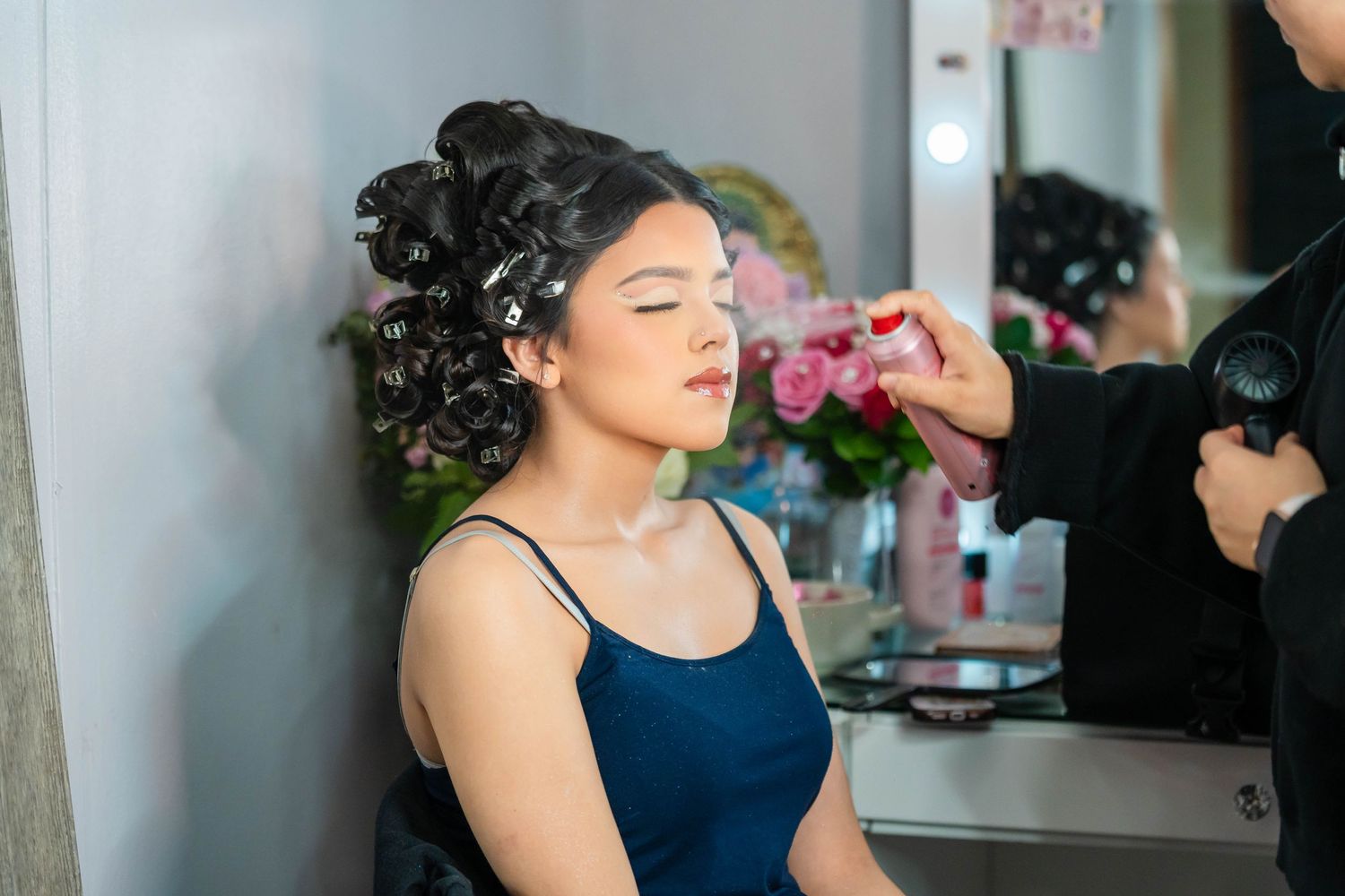 A bride in a navy blue dress sits in a salon chair getting her hair styled for a special occasion.