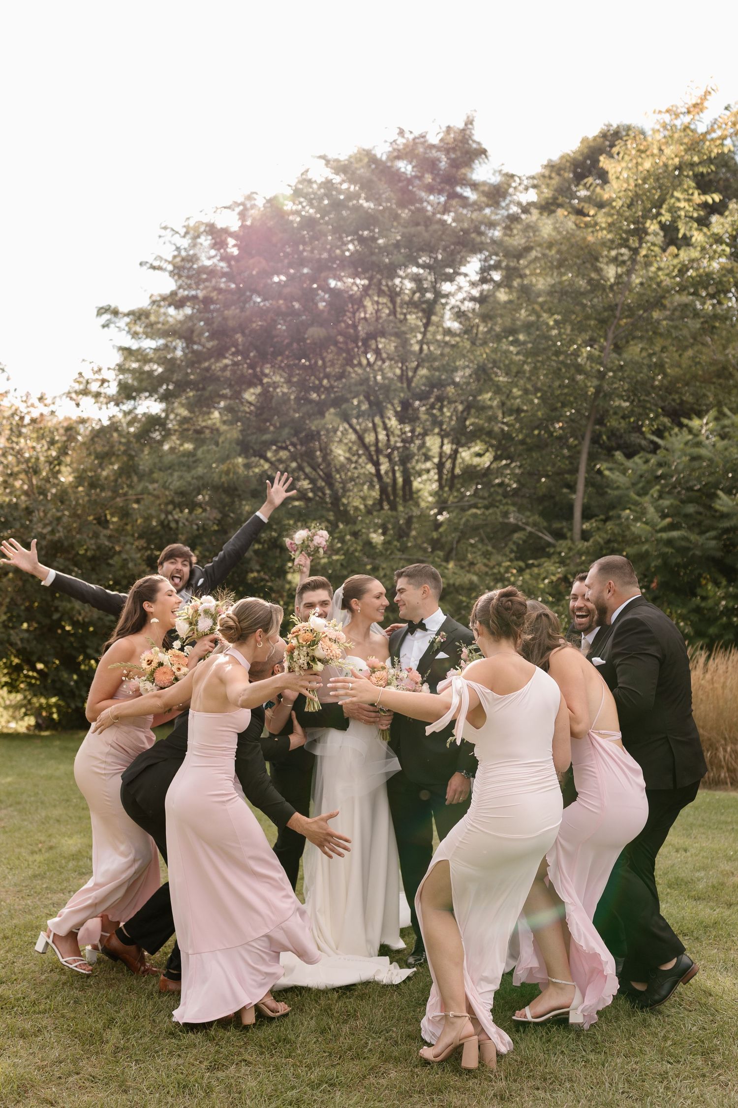 Wedding party in blush pink dresses and black suits playfully pose together in a garden setting at sunset.