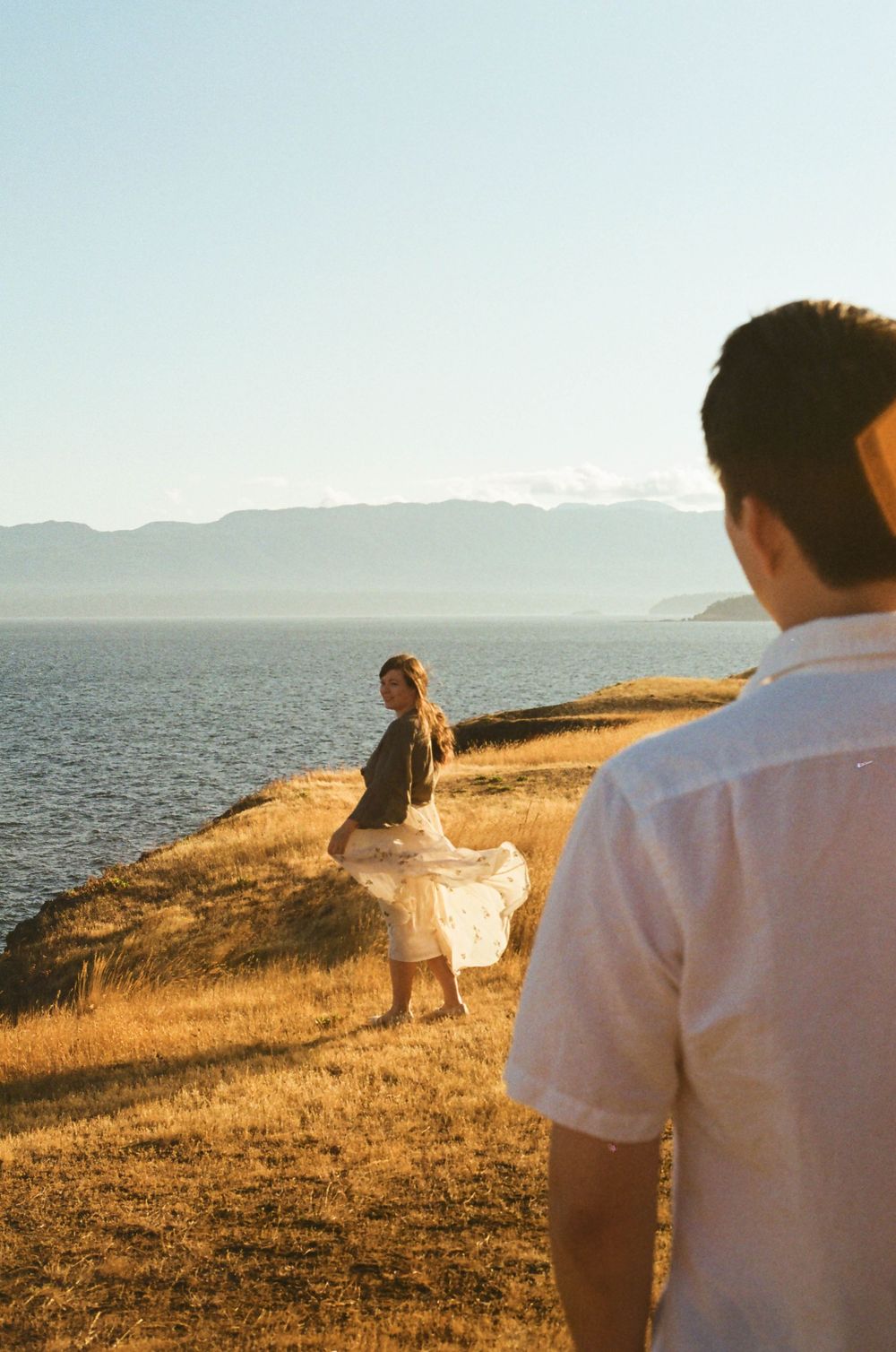 Two people stand on a scenic coastal cliff at golden hour as sunlight bathes the grassy landscape in warm tones.