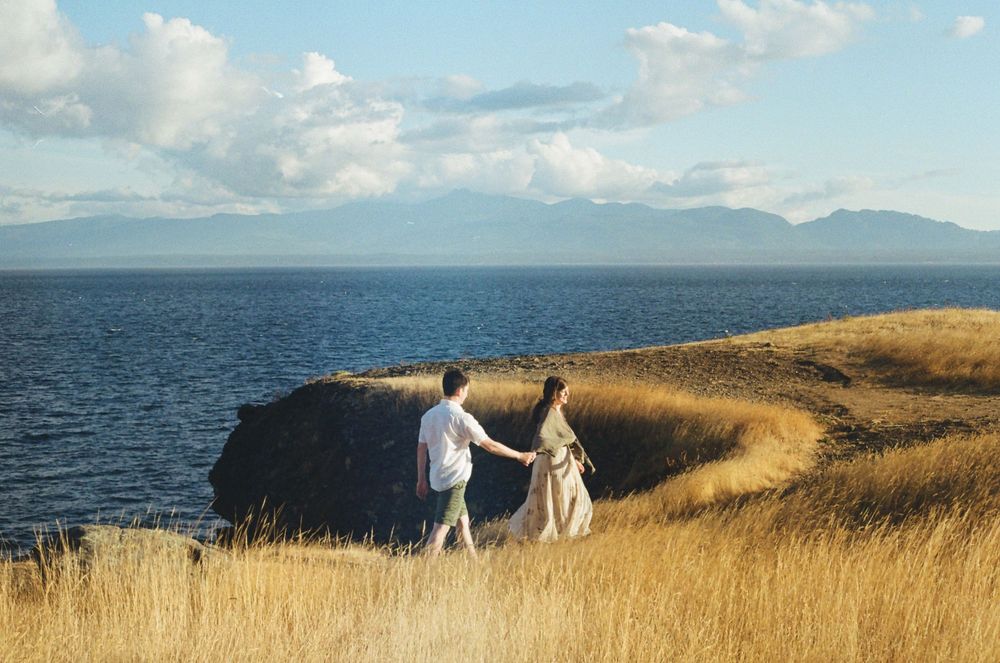 Wide landscape view of coastal bluffs with dry grass and ocean meeting distant mountains under blue skies.