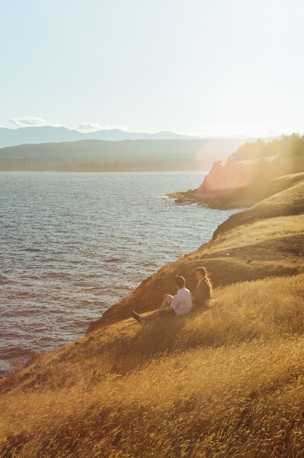 Silhouettes sit on golden grass overlooking a serene mountain lake at sunset with sunlit cliffs in the distance.