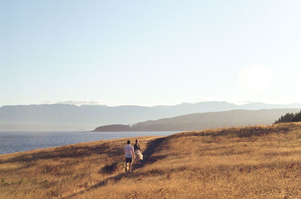 Hikers traverse a golden hillside path with distant mountains and ocean views under a bright blue sky.