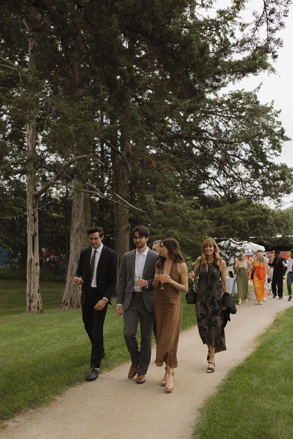 Wedding guests walking along tree-lined path in formal attire.