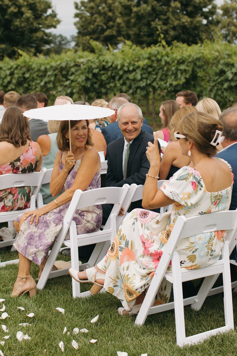 Wedding guests in floral dresses laugh together while seated in white chairs on a grassy lawn.