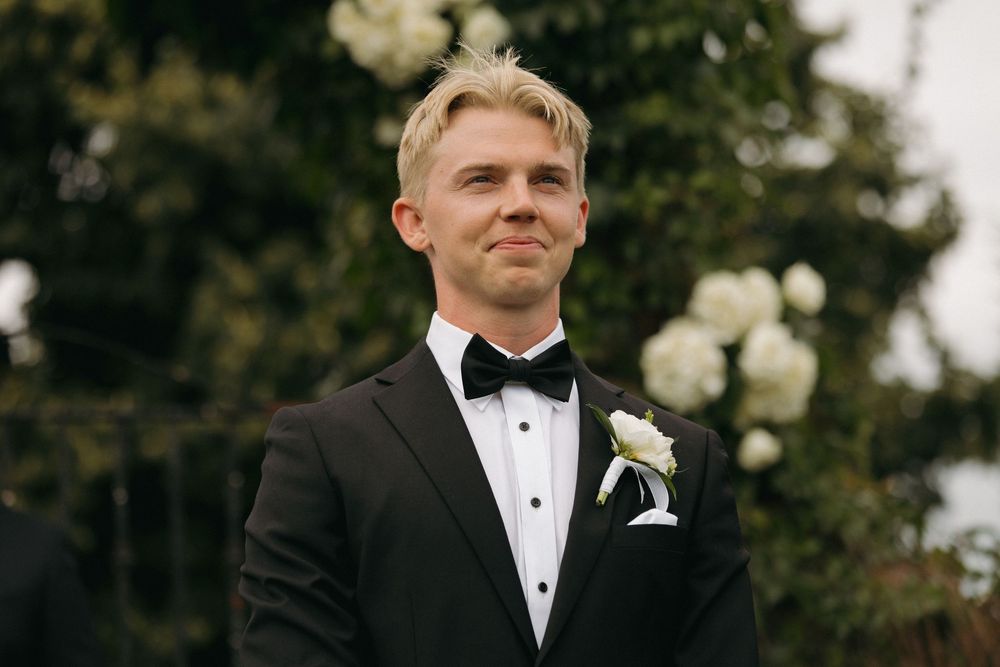 Groomsmen in black tuxedos and bow ties stand together at an outdoor wedding ceremony.