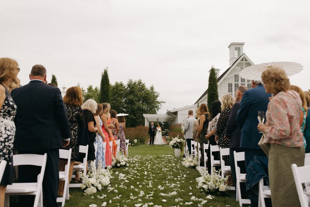 White folding chairs line a grassy outdoor wedding aisle decorated with scattered flower petals.