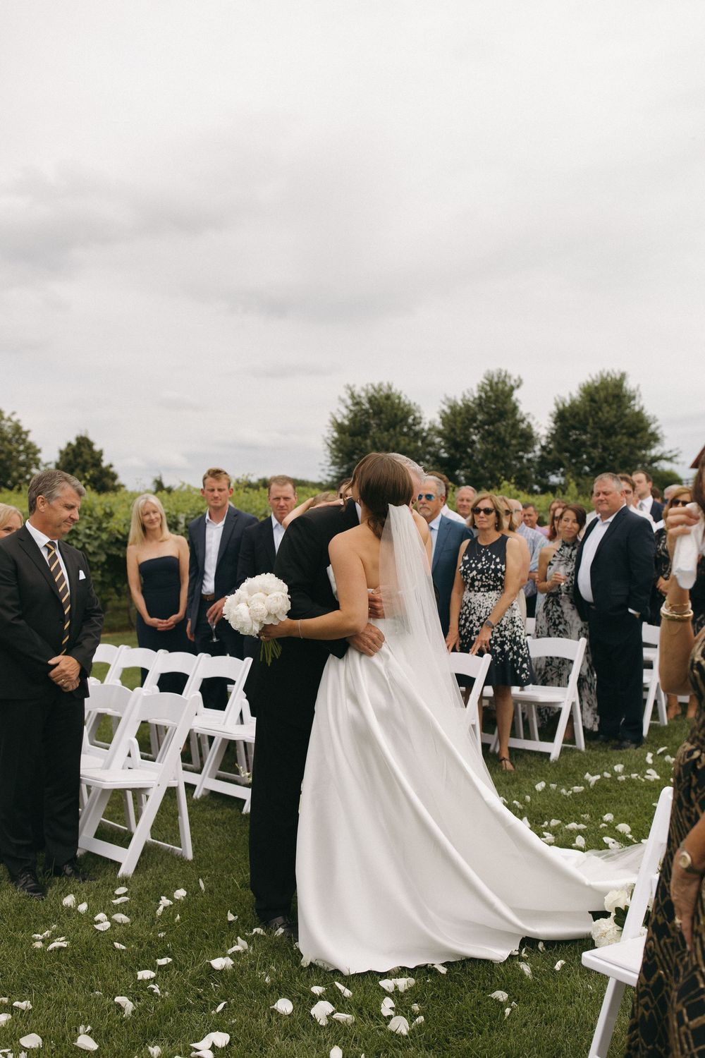 Wedding ceremony guests gather on a lawn with white chairs and scattered petals.
