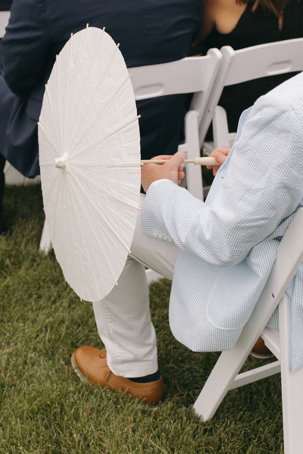 Person in seersucker suit holds white parasol while seated in white folding chair.