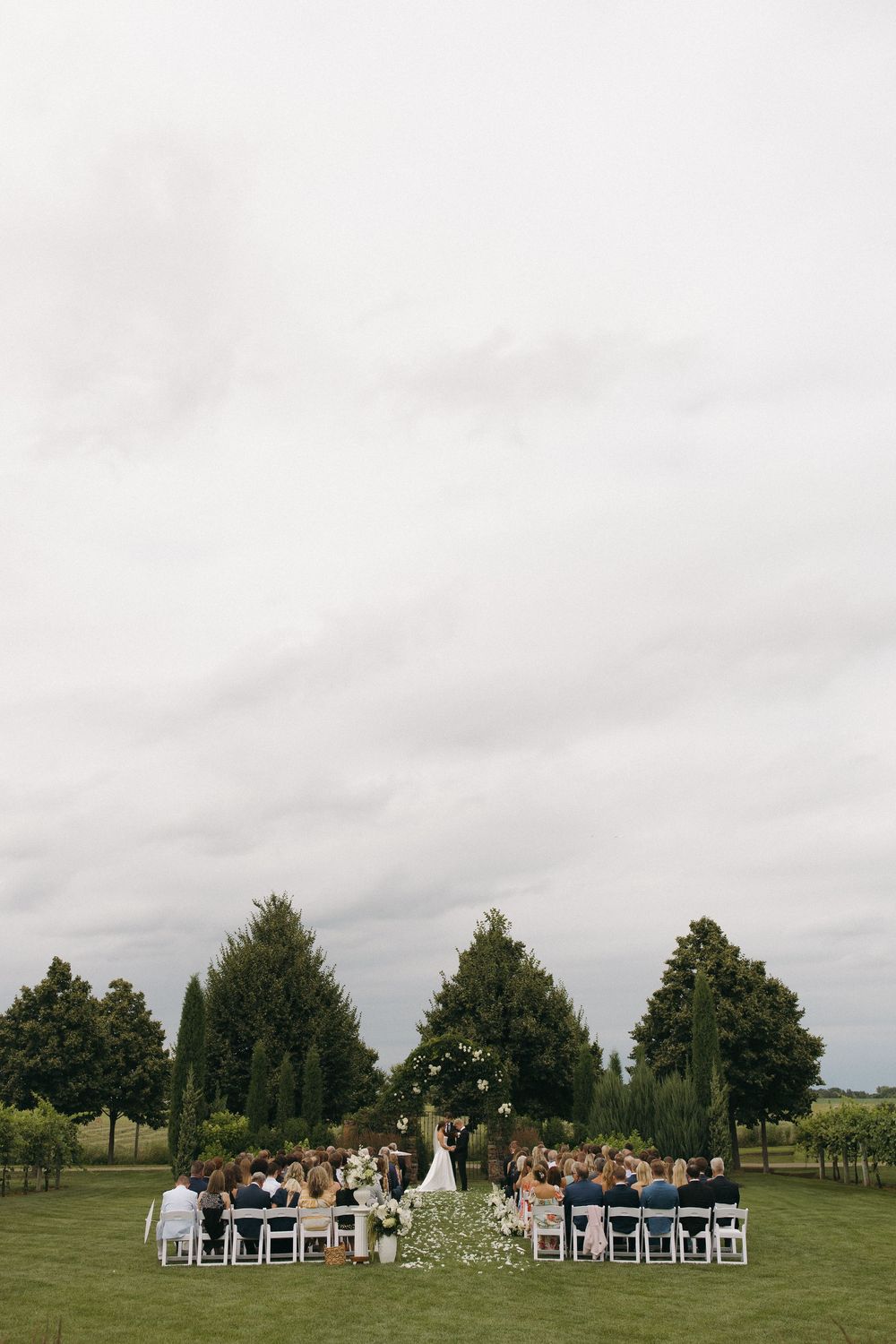 Distant view of outdoor wedding ceremony with white chairs arranged on sprawling green lawn.