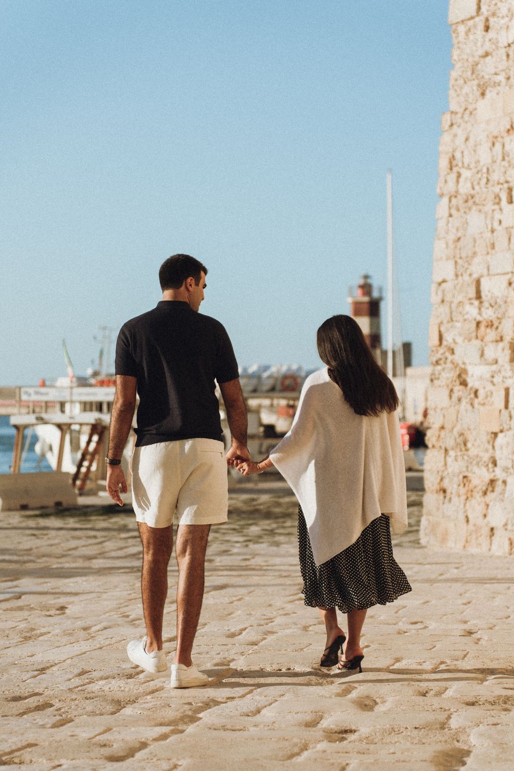Couple walking along a stone seaside path during a sunny day, backdropped by blue ocean views.