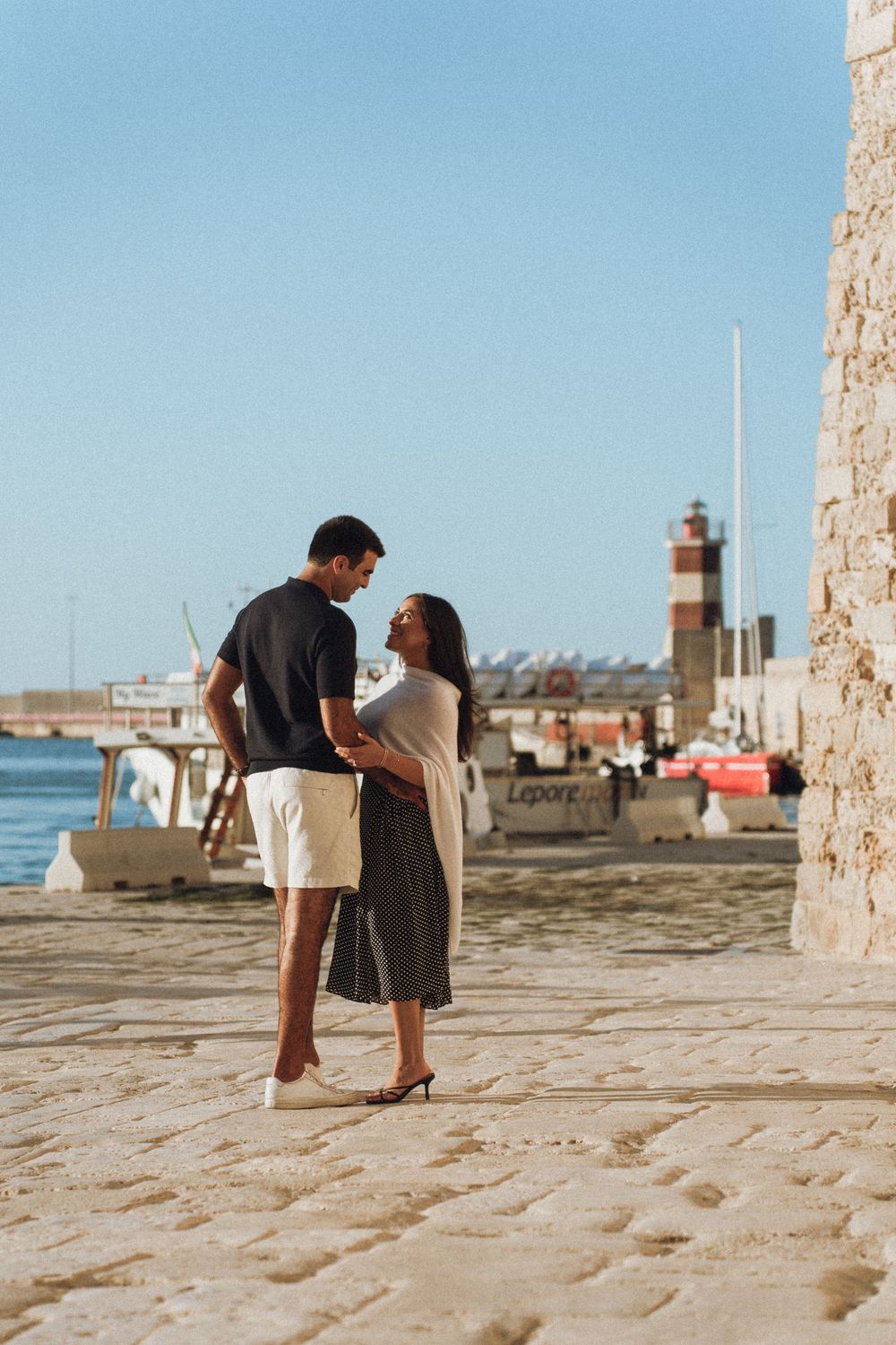 A playful couples photoshoot by the harbor with stone pavement and blue skies in the background.