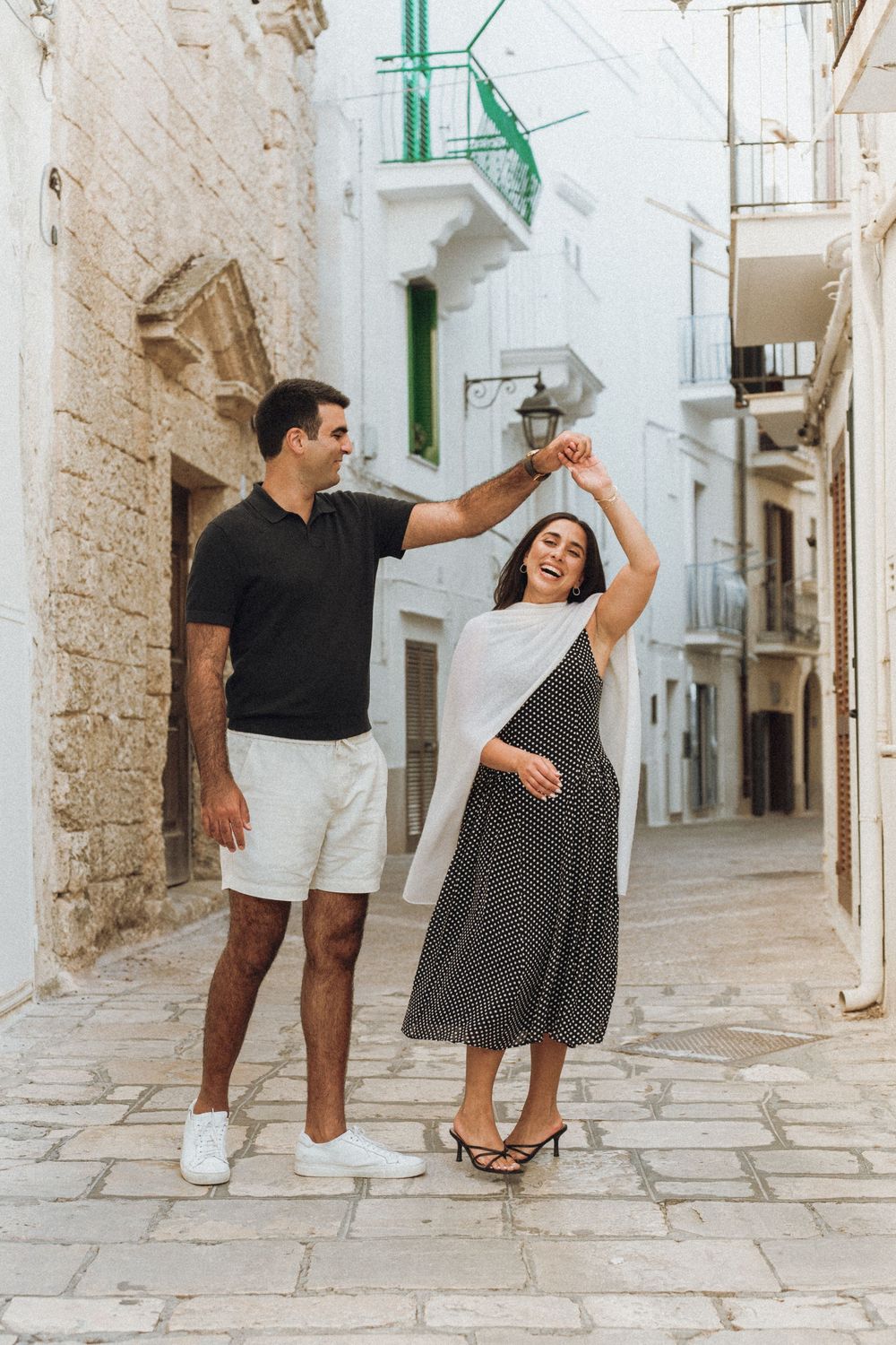 A couple dancing together on a charming cobblestone street in a Mediterranean town with white walls and green accents.