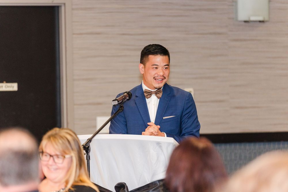 Business professionals seated in an audience at a corporate conference listen to a presenter in a blue suit.