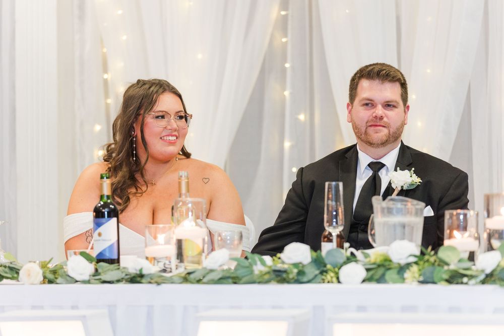 Wedding party seated at a long table with white linens and decor during a reception celebration.