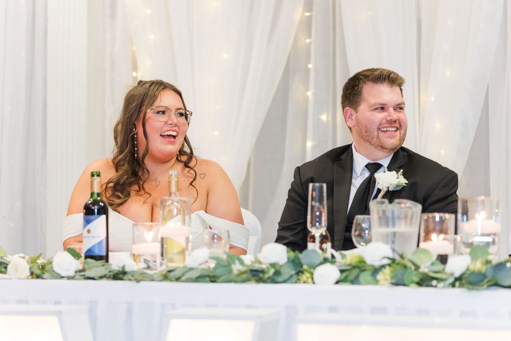 Wedding party seated at head table during reception with decorative floral centerpieces and white drapes in background.
