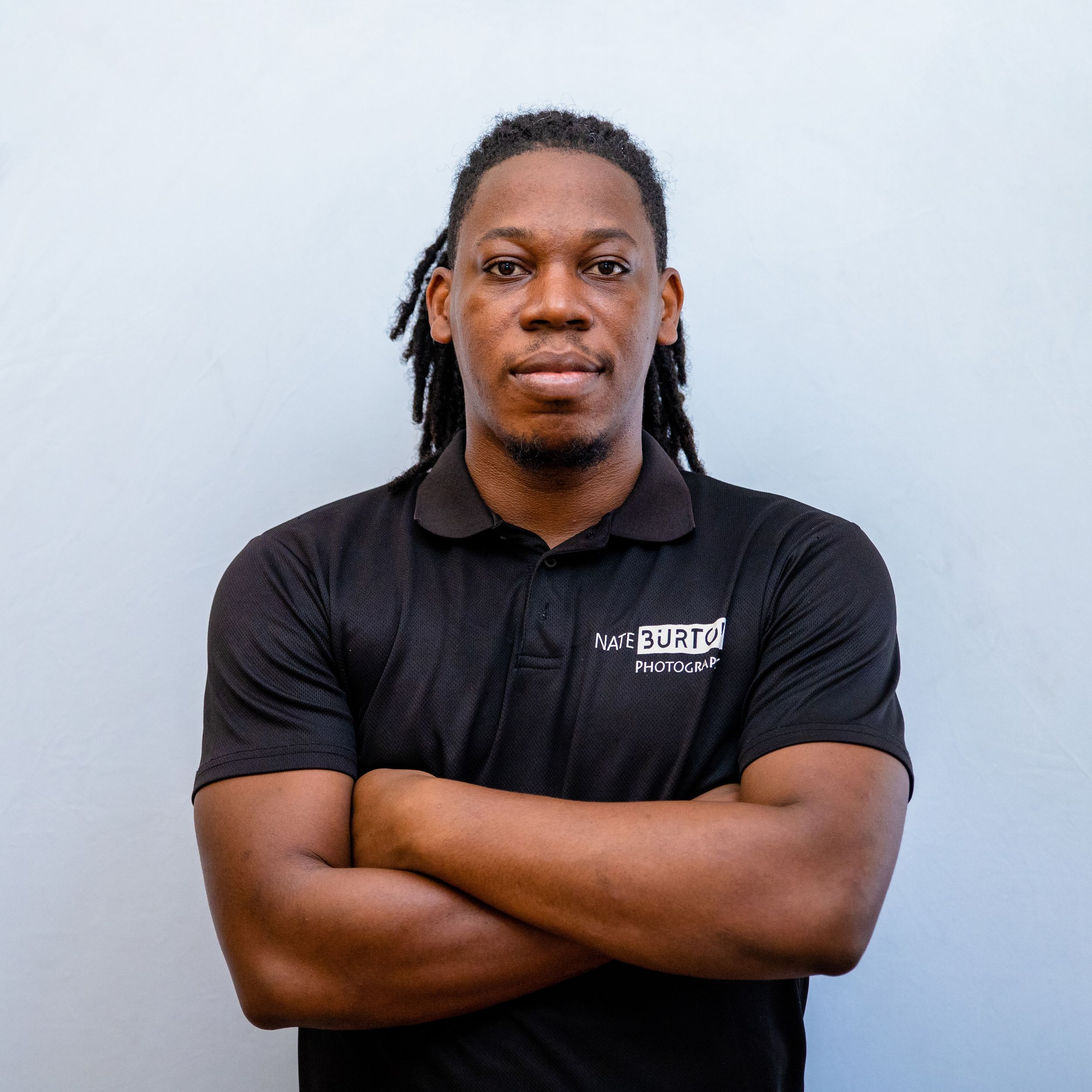 Professional portrait of a person wearing a black polo shirt with dreadlocks against a grey background.