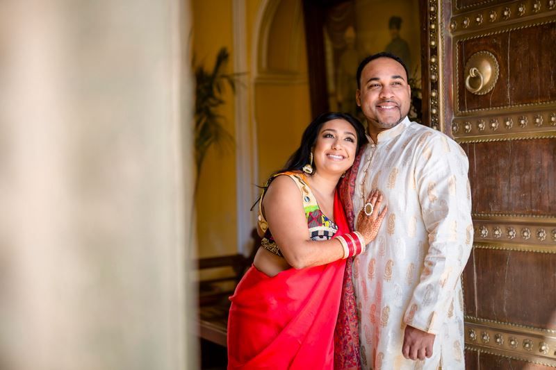A couple in traditional Indian attire poses together in front of an ornate wooden door in a warm-lit hallway.