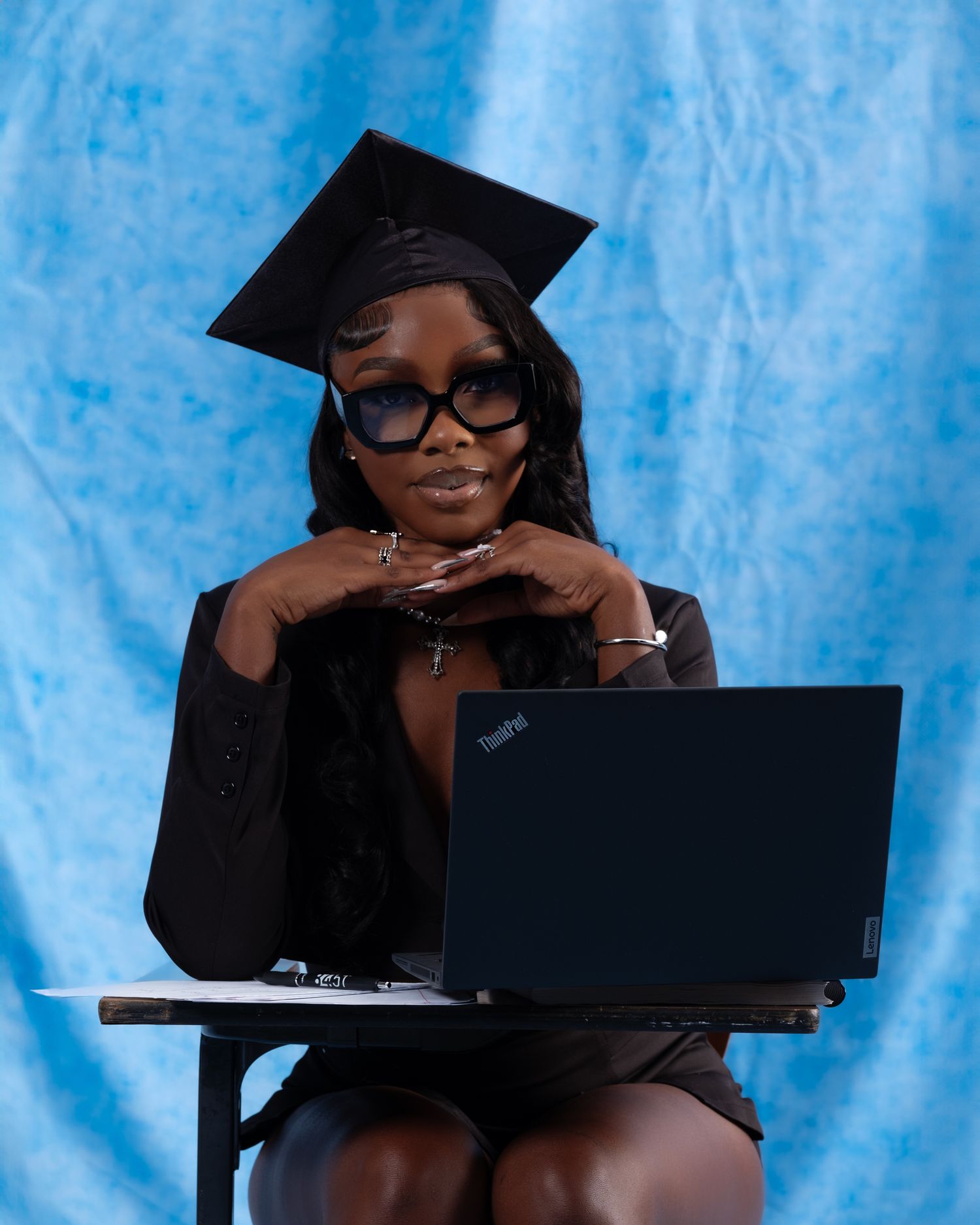Graduate in cap and gown poses with laptop against blue backdrop.